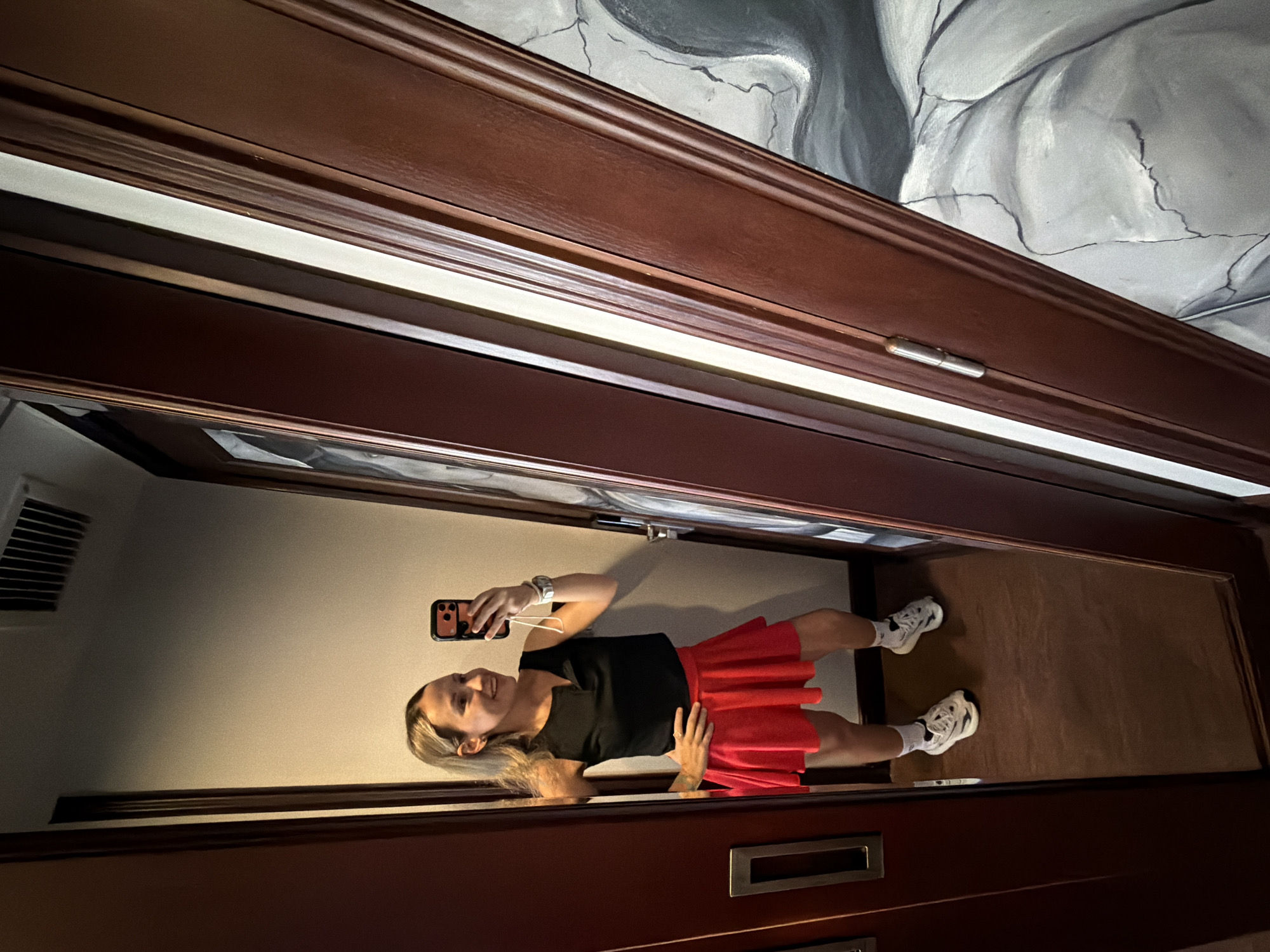 A young Caucasian woman with blonde hair pulled back in a ponytail is standing in front of a mirror and taking a selfie with her phone. She is wearing a black sleeveless tank top and a red pleated skirt, white socks, and athletic shoes. Her left hand is on her hip, and she is smiling at her reflection. The mirror is large and appears to be in a room with dark wood trim and a brown carpeted floor. To the right of the mirror is a door with a painting on it. The overall impression is of a casual, personal moment captured in a hotel room or dressing area. This image was taken in Thuận Hóa Ward, Vietnam.