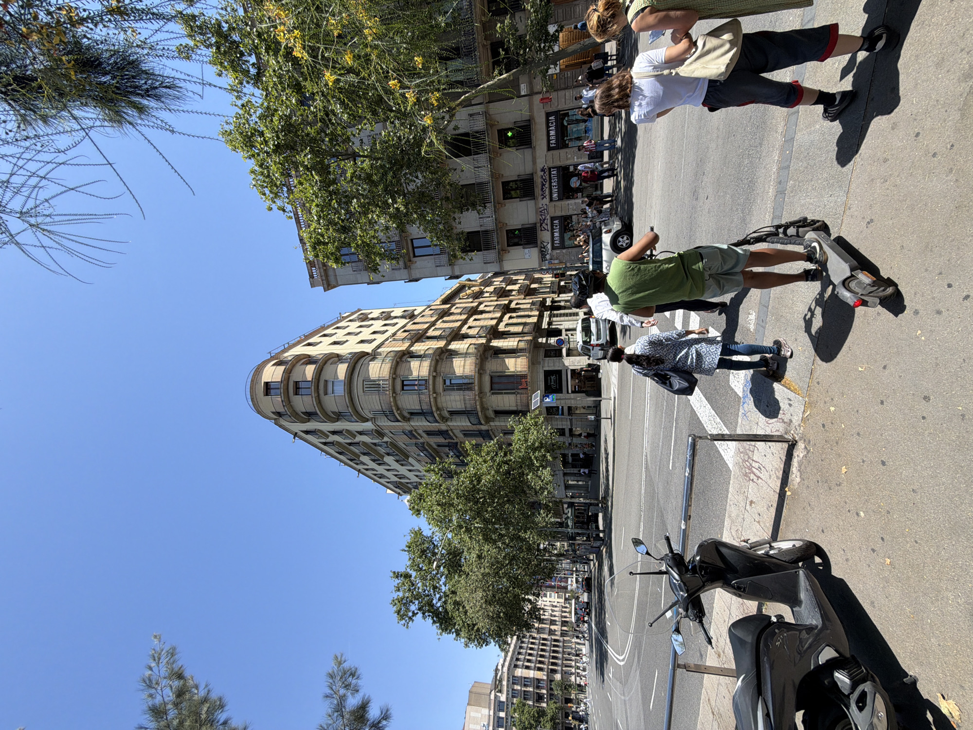 The image captures a street scene in Barcelona, Spain, likely on a sunny day. In the foreground, a dark gray scooter is parked on the sidewalk. A person on a gray electric scooter is moving across the street, wearing a green sleeveless shirt and light blue shorts. To their right, two individuals are standing on the sidewalk. One person, facing away, wears a white t-shirt and dark shorts with red trim, carrying a light-colored tote bag. Another person, partially visible on the far right, wears a green patterned dress and also carries a bag.
Behind them, the street is a busy intersection with vehicles and pedestrians. The buildings in the background are typical of Barcelona architecture, with ornate balconies and light-colored facades. One prominent building is a rounded corner structure with multiple stories and balconies. Several trees with lush green foliage are scattered throughout the scene, providing shade. The sky is clear and blue. Signs for "FARMACIA" and "UNIVERSITAT" are visible on one of the buildings. The overall impression is a vibrant urban environment on a bright day.