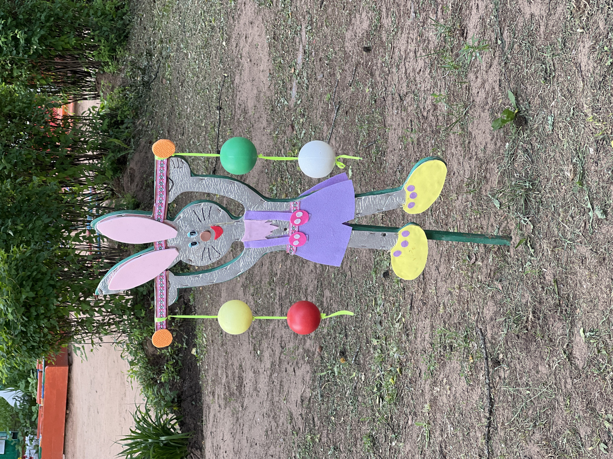 The image shows a wooden cutout of a gray bunny in a park. The bunny is wearing a purple dress with pink straps and yellow shoes. It is holding a bar across its chest, from which hang four colorful balls: yellow, red, green, and white. The bunny has pink ears, blue eyes, a red mouth, and whiskers. The background consists of dirt, grass, and green bushes. In the upper left corner, there are orange rectangular structures. The image is taken from a slightly low angle, looking up at the bunny. The overall scene depicts an outdoor recreational area, possibly a playground or a decorative element in a park. The location is identified as Советско-Никольское, Russia.