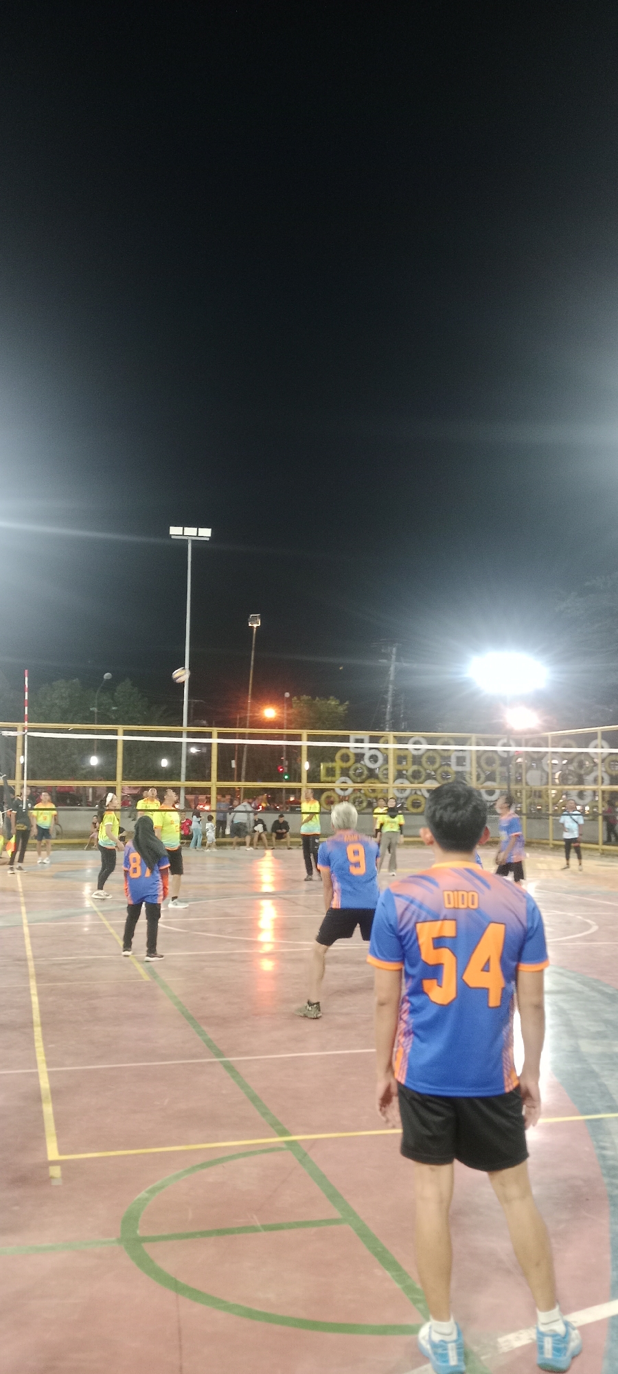 In Tuban, Indonesia, at night, a group of people are playing volleyball on an outdoor court illuminated by floodlights. The court has red and tan colored flooring with green and yellow lines marking the boundaries. A volleyball net is set up across the middle of the court. In the foreground, facing away from the camera, a person wearing a blue and orange jersey with the number "54" and the name "DIDO" on the back is visible. Another person in the middle ground, also wearing a blue and orange jersey, has the number "9" on their back. Several other players in similar colored jerseys, predominantly yellow and blue, are scattered across the court, engaged in the game. Spectators can be seen in the background, some sitting and others standing. Trees and poles with lights are visible beyond the court fence. The overall atmosphere suggests a casual, evening sporting event.