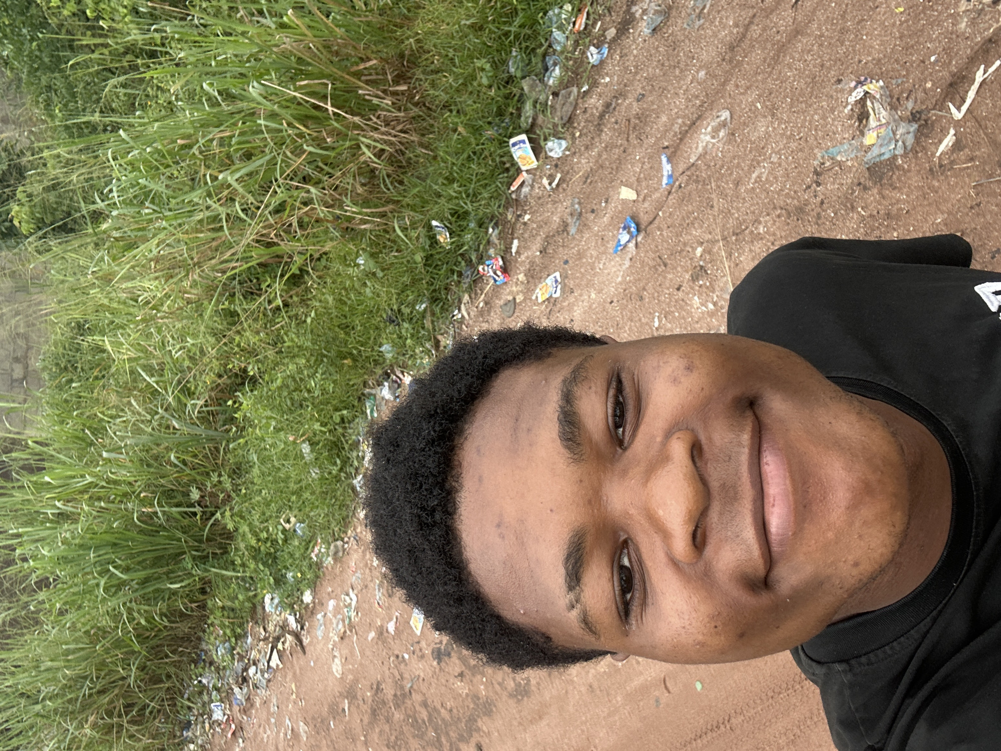A young Black male, likely a teenager, smiles directly at the camera in a selfie. He is wearing a black t-shirt. The background suggests an outdoor, somewhat unkempt environment. To the left and behind him, tall green grass grows abundantly. The ground in front of him and to his right is a reddish-brown dirt path, littered with various pieces of trash, including plastic wrappers, bottles, and other debris. A rough, light-colored wall is visible in the far background behind the grass. The overall scene conveys a sense of an informal, possibly rural or suburban, setting within Ihiagwa, Nigeria.