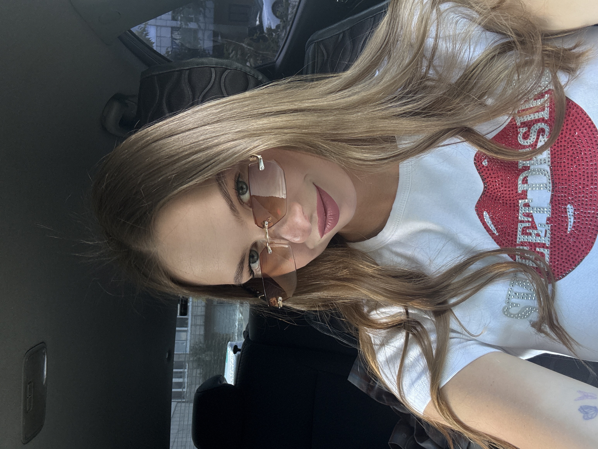 The media file is a close-up, rotated selfie of a young woman sitting in a car. She has long, blonde hair that falls over her shoulders and a white t-shirt adorned with a large, red, sequined lip graphic and text. She is wearing stylish, rimless sunglasses with a warm-toned tint and has rosy-pink lipstick on. Her expression is relaxed, with a slight smile. The background shows the interior of a car, including a black headrest and part of a seat. Through the car window, a building with multiple floors and windows is visible, suggesting an urban or suburban environment. The lighting indicates it's daytime. The overall mood is casual and confident. The text on the t-shirt is partially obscured but appears to read "DIJSS" in silver rhinestones.