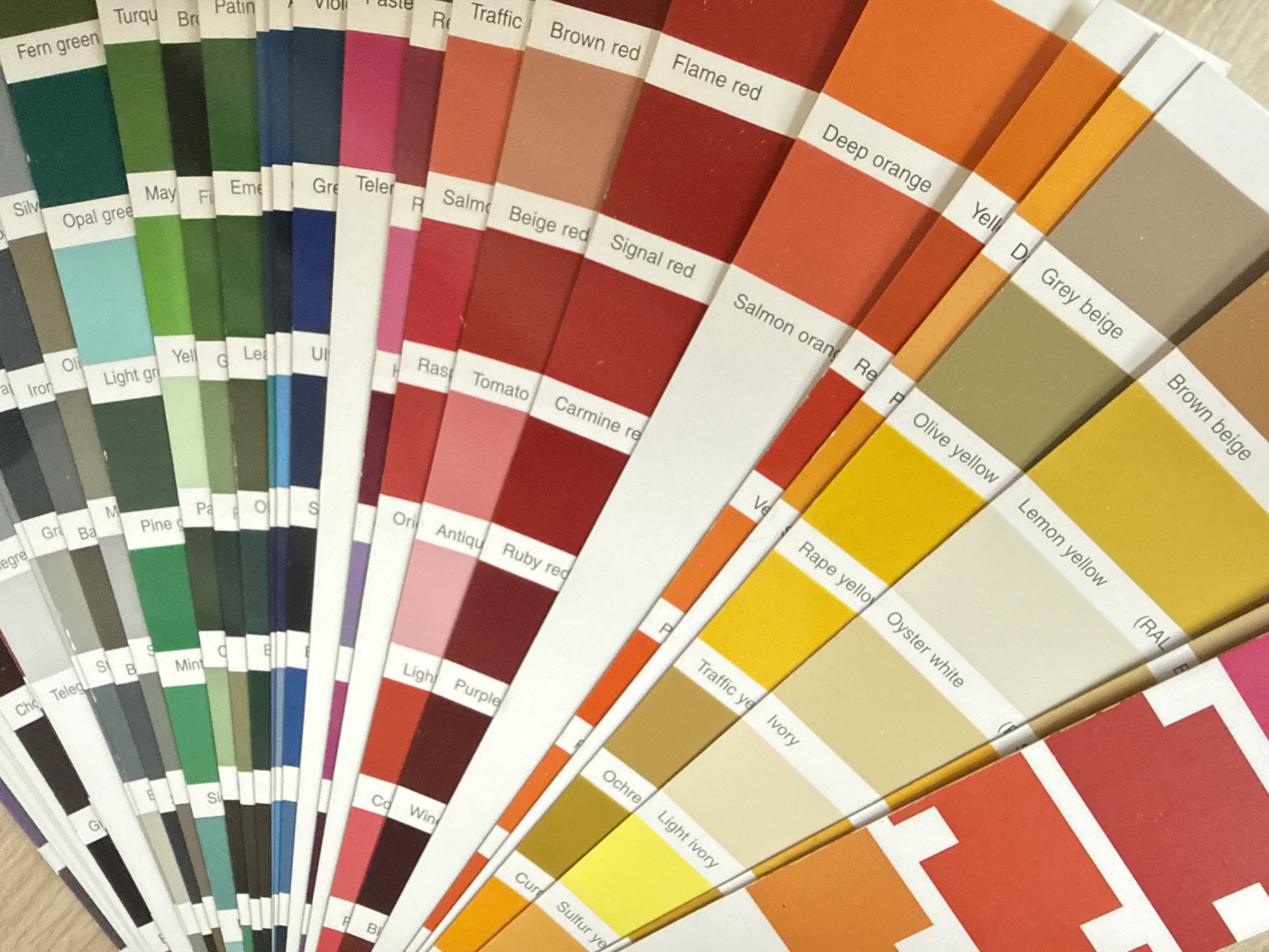 The image displays a color swatch fan, likely a RAL color system chart, fanned out on a light brown, textured surface. The swatches are arranged in a semi-circular pattern, revealing a spectrum of colors from greens and blues at the top to reds, oranges, and yellows at the bottom. Each color swatch has a white label strip with text indicating the color name and possibly a code. Some of the visible color names include "Fern green", "Opal gree", "May F", "Mint", "Light Purple", "Ruby red", "Carmine re", "Signal red", "Salmon orang", "Deep orange", "Ochre", "Light ivory", "Oyster white", "Lemon yellow", and "Brown beige". The lighting suggests a close-up shot, with a shallow depth of field, making the foreground swatches sharp and the background slightly blurred. There are no people, animals, or specific landmarks visible in the image. The context of Stara Zagora, Bulgaria is not discernible from the visual information provided in the photograph.