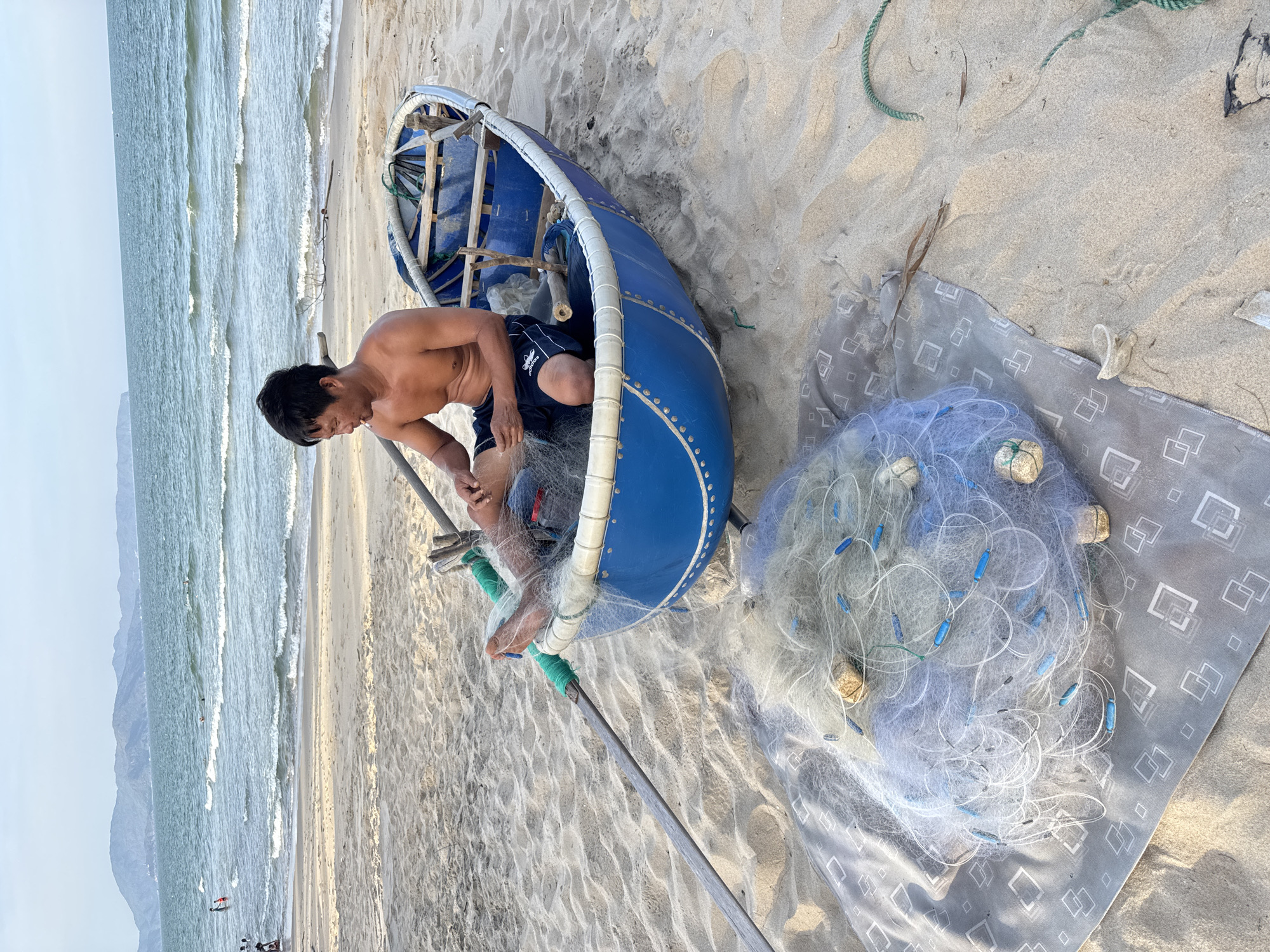 A shirtless man with dark hair sits in a blue, round boat on a sandy beach in South Quy Nhon Ward, Vietnam. He is wearing dark shorts and appears to be untangling a fishing net. Beside the boat, on a grey patterned cloth, lies another bundle of fishing net. In the background, the ocean stretches towards a hazy coastline with mountains. Waves are breaking on the shore, and a few people are visible in the water and on the beach further away. The scene depicts a tranquil moment of preparation or post-fishing activity.