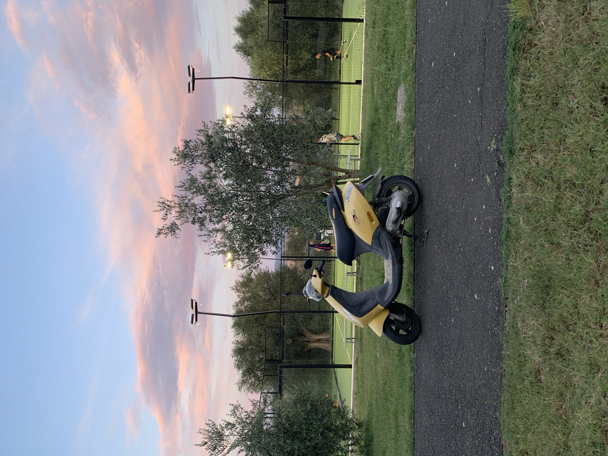 The image depicts a yellow scooter parked on a paved path adjacent to a grassy area. Beyond the grass and path, a fenced-off green court, resembling a paddle tennis court, is visible. Two individuals are on the court, one appearing to be playing and the other observing. Olive trees flank the court on both sides, and a tall light pole with multiple lamps stands near the center of the court. The sky above is a gradient of blue, with pink and orange clouds, suggesting it is either sunrise or sunset. The location is identified as San Quirico d'Orcia, Italy.