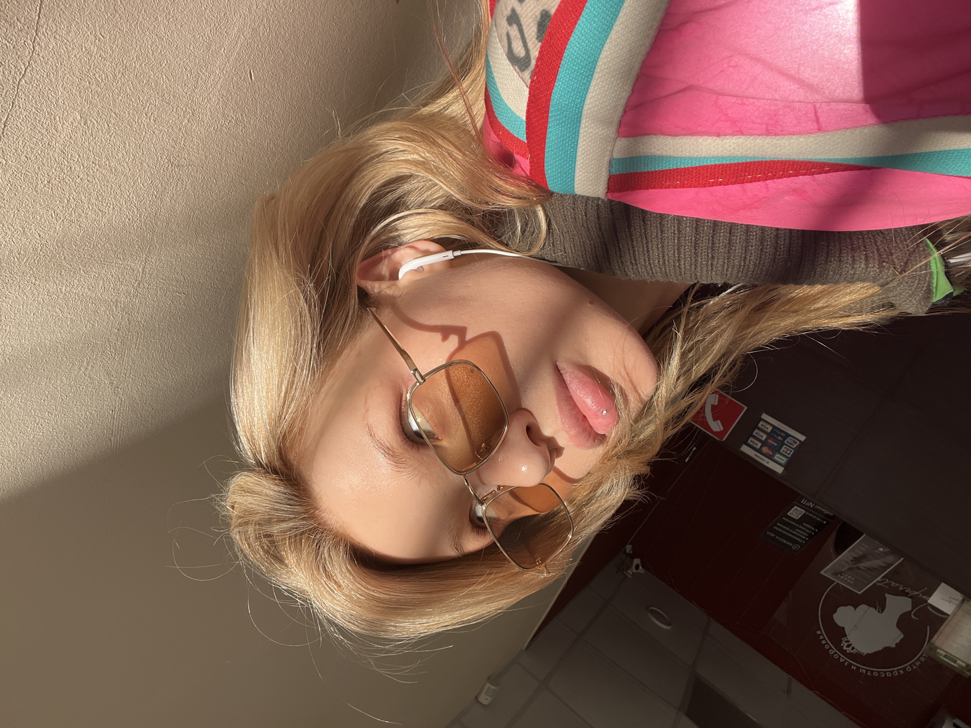 The image depicts a close-up, upward-angled indoor shot of a blonde individual, likely female, positioned within a reception area. This location is identified as the "Центр Красоты и Здоровья Императрица" (Empress Beauty and Health Center) at ул. Ленина, 58, Chaikovsky, Perm Krai, Russia.
The subject's face is partially illuminated by direct sunlight from the upper left, creating distinct highlights and shadows. She wears aviator-style sunglasses with amber-tinted lenses and thin gold-colored frames. A small, clear stud lip piercing is visible on her upper lip, slightly right of the philtrum. A white wired earbud is present in her right ear. She is wearing a dark olive green or brown ribbed sweater. A vibrant pink bag with white, light blue, and red stripes is visible in the lower left foreground.
The background to the subject's left features a dark red/maroon wall or counter. Mounted on this surface is a white circular logo displaying the silhouette of a woman's head with a crown, encircled by the Russian text "ЦЕНТР КРАСОТЫ И ЗДОРОВЬЯ" (Beauty and Health Center). Below this, partially obscured text confirms "Императрица" (Empress). Further down, signs displaying payment card logos (VISA, MasterCard, Mir) and QR codes are visible, along with a red emergency phone symbol. A small white security camera is affixed to this wall near the ceiling. The ceiling above features standard white tiles and recessed lighting. To the subject's right, a textured beige wall is visible, with a portion strongly lit by sunlight and another section in shadow.
