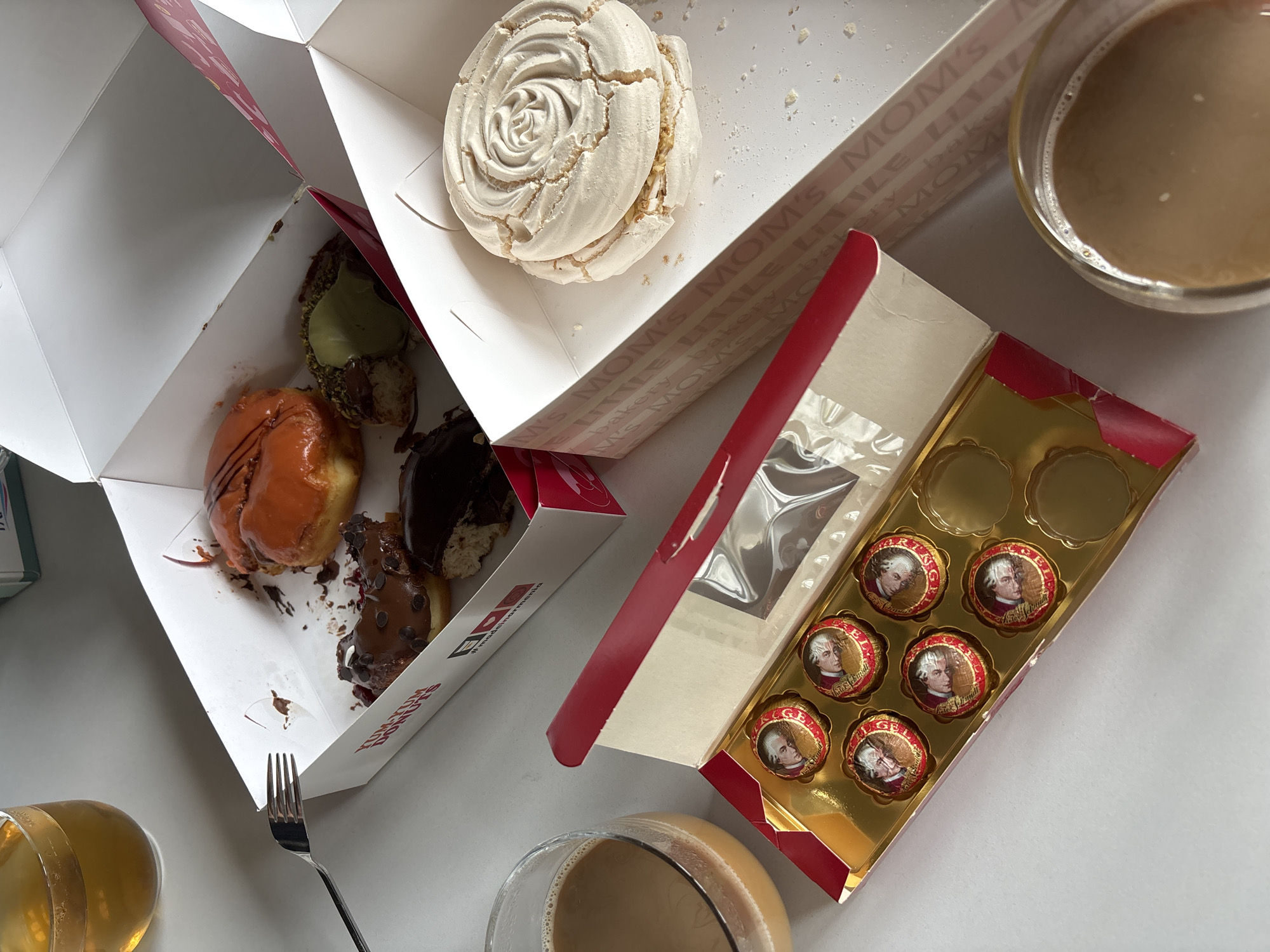 The image shows an overhead shot of a food arrangement. It includes a box of four doughnuts, a meringue pastry in a box labeled "Mom's Little Bakery," and a box of wrapped Mozart Kugeln chocolates with a red and gold design. Also visible are two glasses of milky coffee and a fork. The doughnut box is labeled "Dunkin' Donuts." The environment suggests a casual setting, possibly a table top. Given the Dunkin' Donuts reference, the location is likely in the city of Yerevan, Armenia, as it is one of the few cities where that franchise is present in combination with the other products shown.