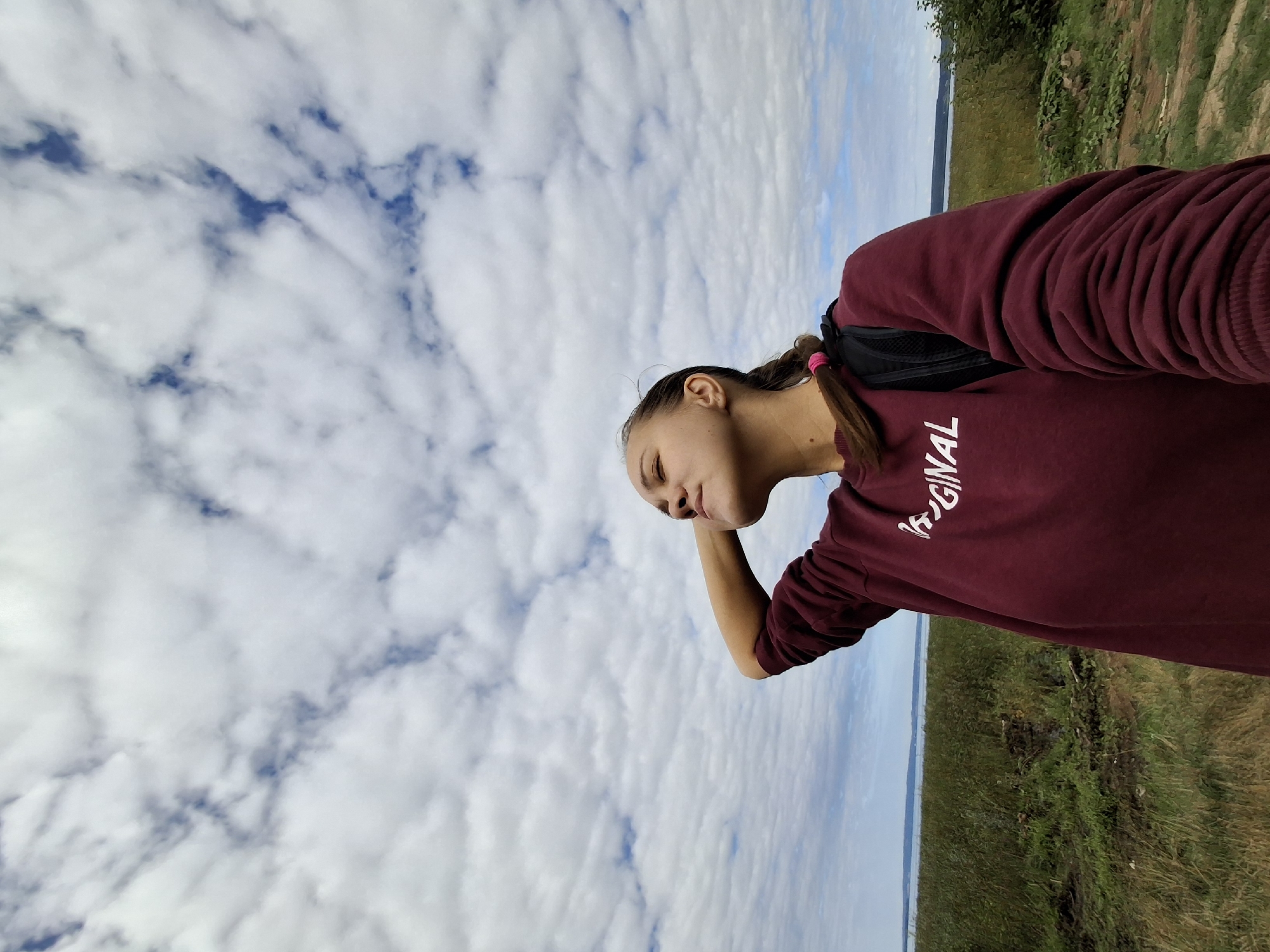 A young woman is taking a selfie outdoors, with her head tilted back and eyes closed as she looks up at a cloudy sky. She is wearing a maroon sweatshirt with white lettering that appears to say "ORIGINAL" and has a black backpack on. Her hair is pulled back in a braid with a pink tie. The background shows a wide expanse of grass and vegetation leading to a body of water, likely a lake or bay, under a vast, cloud-filled sky. The location is identified as Plishkino, Russia.