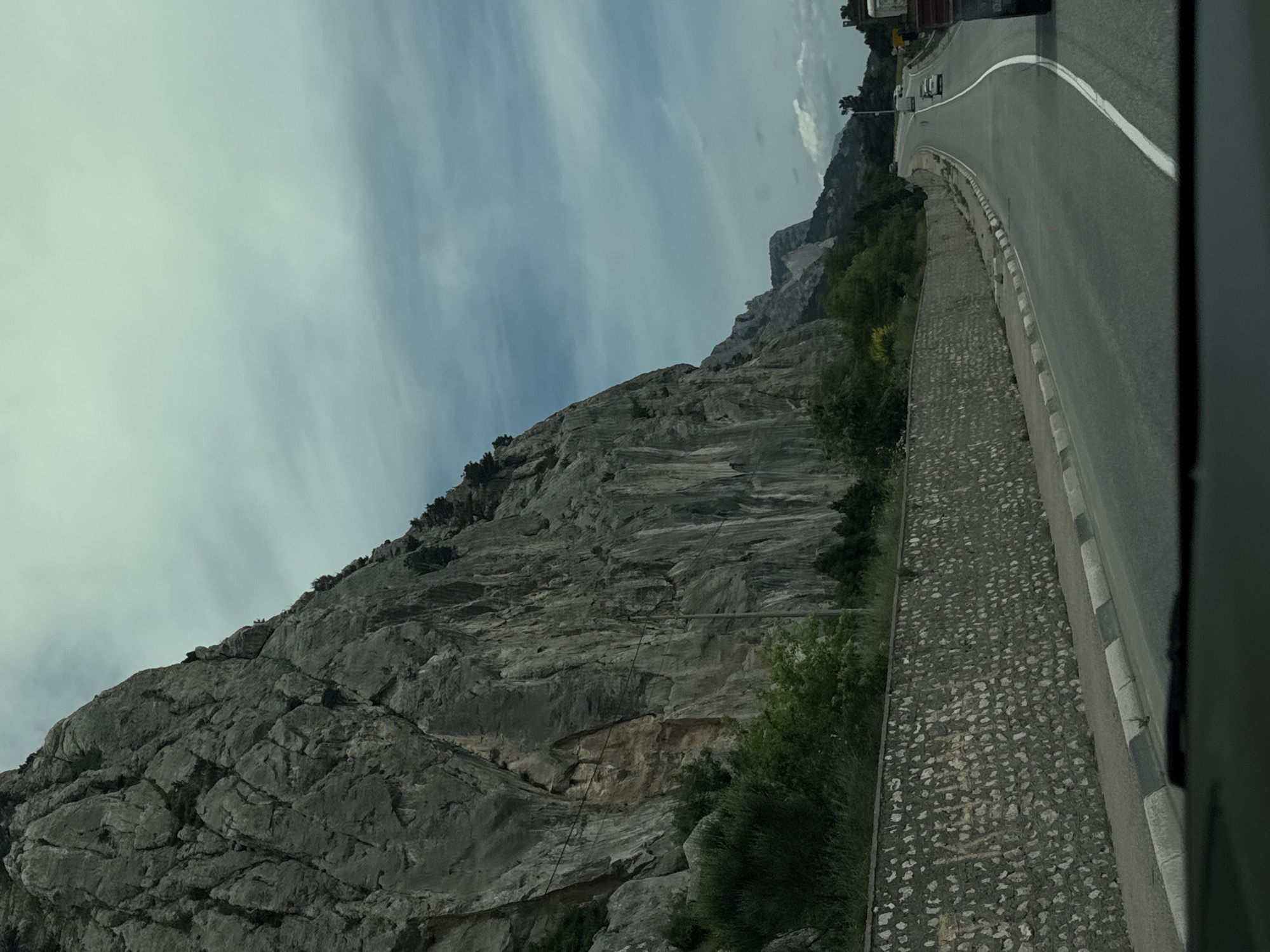 The image presents a landscape featuring a road running alongside a steep, rocky cliff face. The road is paved and marked with a white line, suggesting a two-way traffic flow. Several vehicles are visible on the road. A low, stone barrier separates the road from the cliff edge, with vegetation growing along the base of the cliff. The sky is overcast. The image is taken from a slightly elevated viewpoint, possibly from inside a vehicle. The exact location within Орлиновский округ, Ukraine, is not specified, but the scene suggests a mountainous or hilly region.