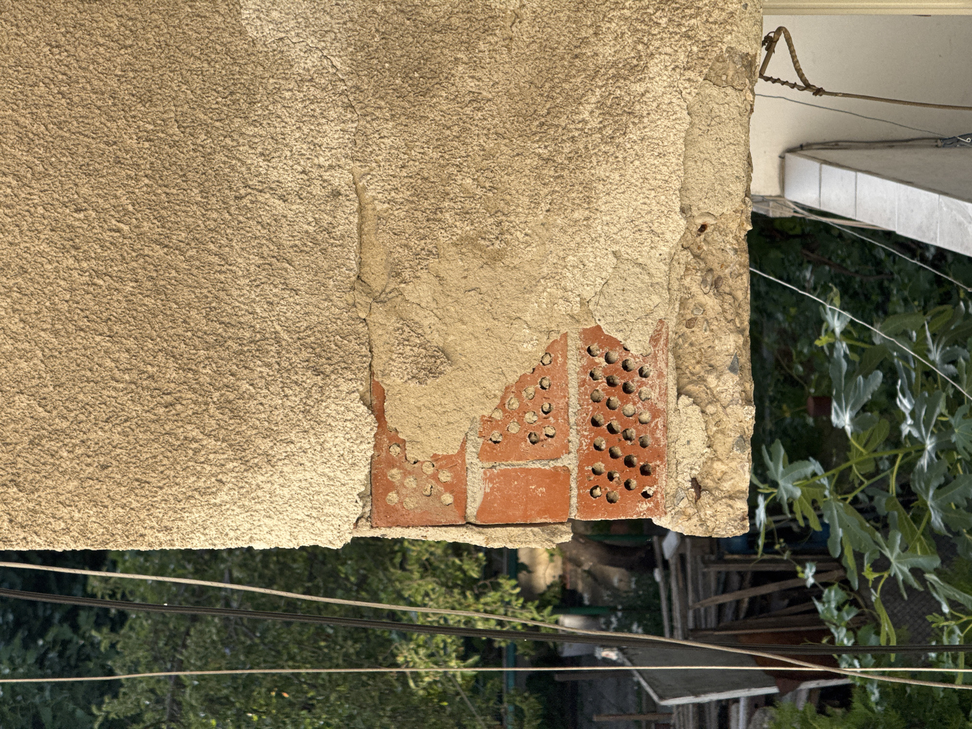 The image captures a close-up view of weathered brickwork and stucco on the exterior of a building. The stucco is peeling away in several areas, revealing the underlying red brick. Some of the bricks have visible perforations, characteristic of certain types of construction material. The scene is set outdoors, with lush green foliage and trees visible in the background, suggesting a natural or garden setting. Several electrical wires or cables run vertically along the left side of the frame. The lower portion of the image shows more of the surrounding environment, including what appears to be a balcony railing and a tiled structure. The overall impression is one of an aging building facade, possibly in a residential area. The provided location information is "city of Sofia, Bulgaria."