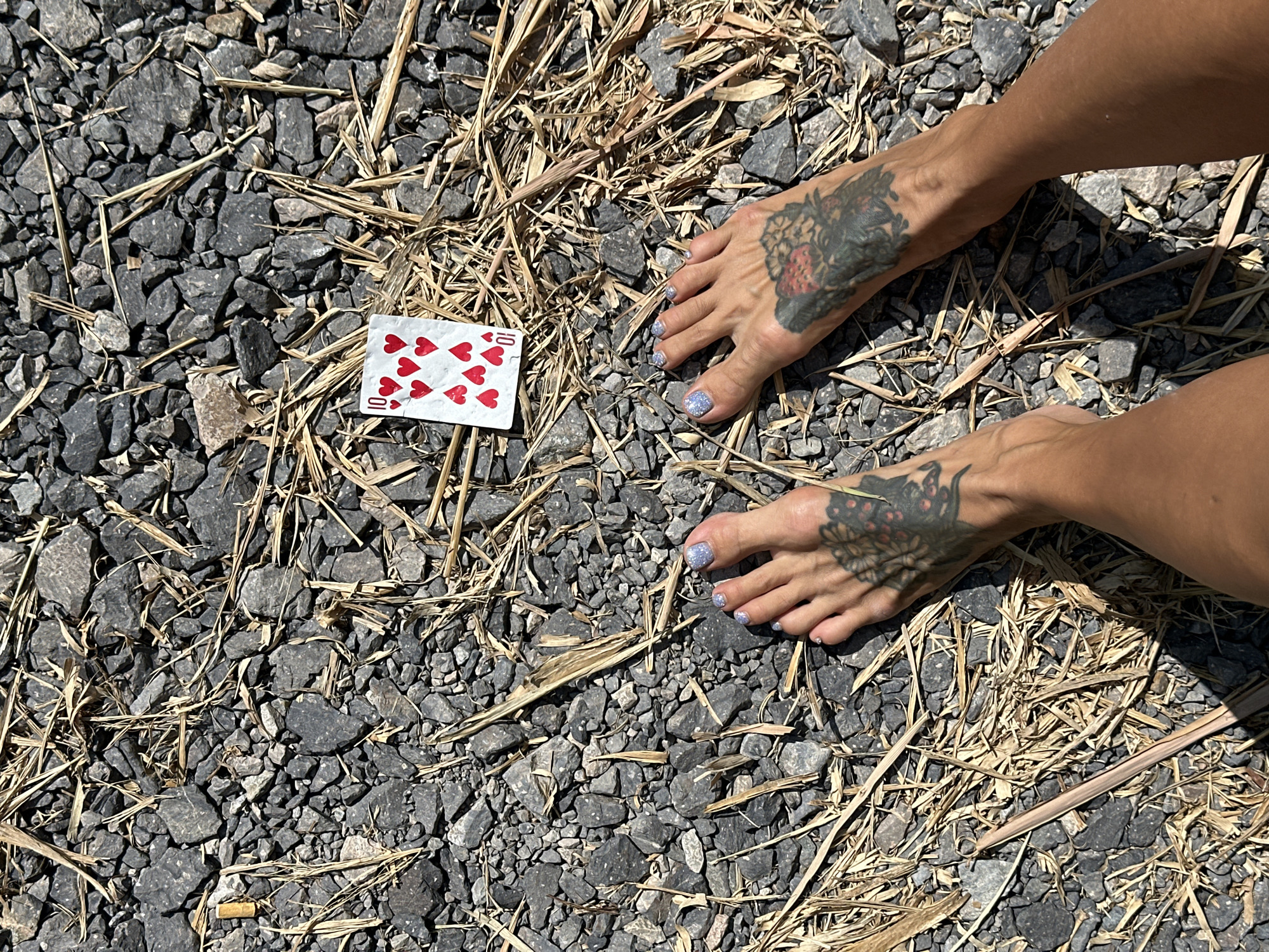 A pair of bare feet, adorned with intricate black tattoos on both ankles, are positioned on a surface of small, grey stones and dry, golden grass. One foot is slightly raised, revealing toenails painted with a sparkling, light blue polish. A single playing card, the ten of hearts, lies face up amidst the stones and grass between the feet. The scene is illuminated by bright sunlight, casting shadows on the ground. The location is the beach in Mũi Né, Vietnam.