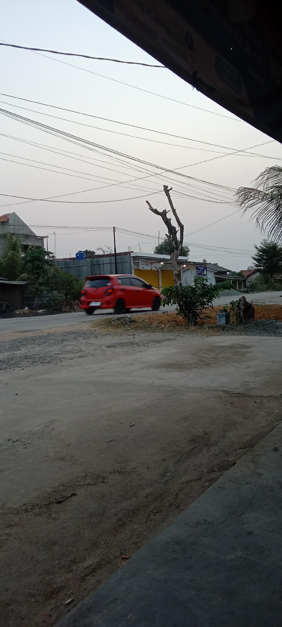 A bright red hatchback car is captured in motion on a paved road, its blurred outline indicating movement. The car is moving from left to right across the frame. In the background, there are several buildings, including a house with a tiled roof on the left and smaller structures that appear to be shops or stalls with yellow awnings. A barren, dead tree with multiple branches stands prominently in the mid-ground, to the right of the car. The sky above is overcast with a pale, diffused light, suggesting either early morning or late afternoon. Utility wires crisscross the sky horizontally. The foreground consists of a rough, unpaved surface and a section of paved ground. The scene appears to be in a semi-urban or rural area.