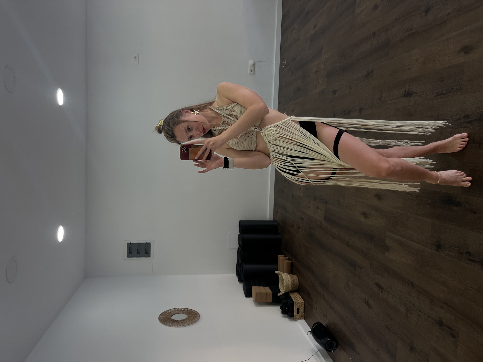 A young woman is taking a selfie in a studio located in Budva, Montenegro. She is wearing a beige crocheted halter top and a fringed skirt over black bottoms. She is holding a red phone in her right hand. She has blonde hair and is wearing gold accessories, including star-shaped earrings and a headband.
The studio has a white wall and a dark wood floor. Near the wall are several black yoga mats stacked next to baskets and small boxes. A round decorative object lies on the floor near the wall.