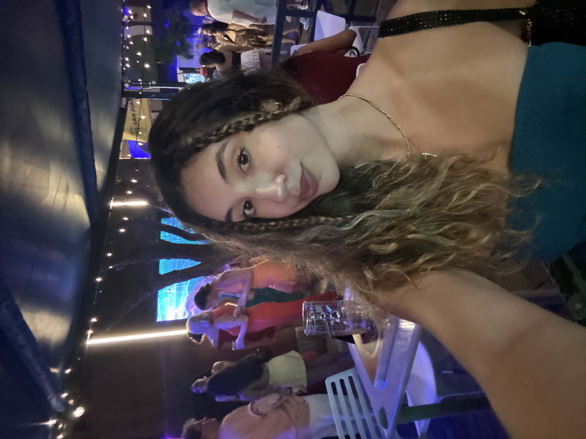 The image shows a young woman taking a selfie with a group of people in the background. The scene appears to be outdoors at night, possibly at a bar or restaurant. The woman is wearing a teal strapless top and a gold necklace. Her hair is long and wavy with some braids. In the background, people are dressed in casual attire, and there are string lights and a screen displaying colorful graphics. A sign with "SAN JUAN" is visible in the upper right portion of the image, indicating the location as San Juan, United States of America. A table with a glass of water is in the foreground.