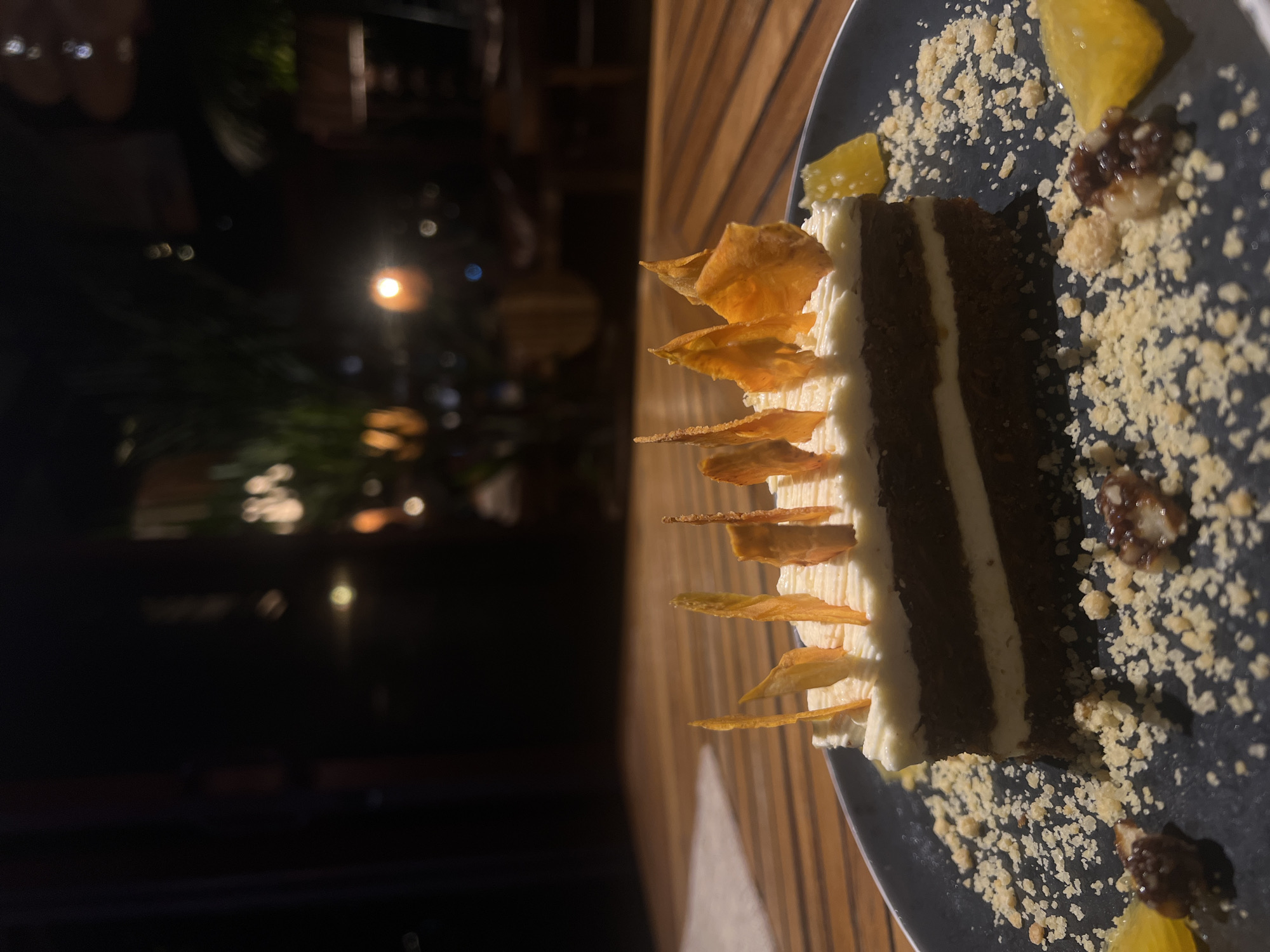 A slice of dark cake with white frosting is plated with candied citrus and crumbled topping. The cake is decorated with thin, orange crisps. It sits on a black plate placed on a wooden surface, possibly a table. The background is dark with faint blurred lights suggesting an outdoor or dimly lit indoor setting. The location is likely North Kuta, Indonesia.