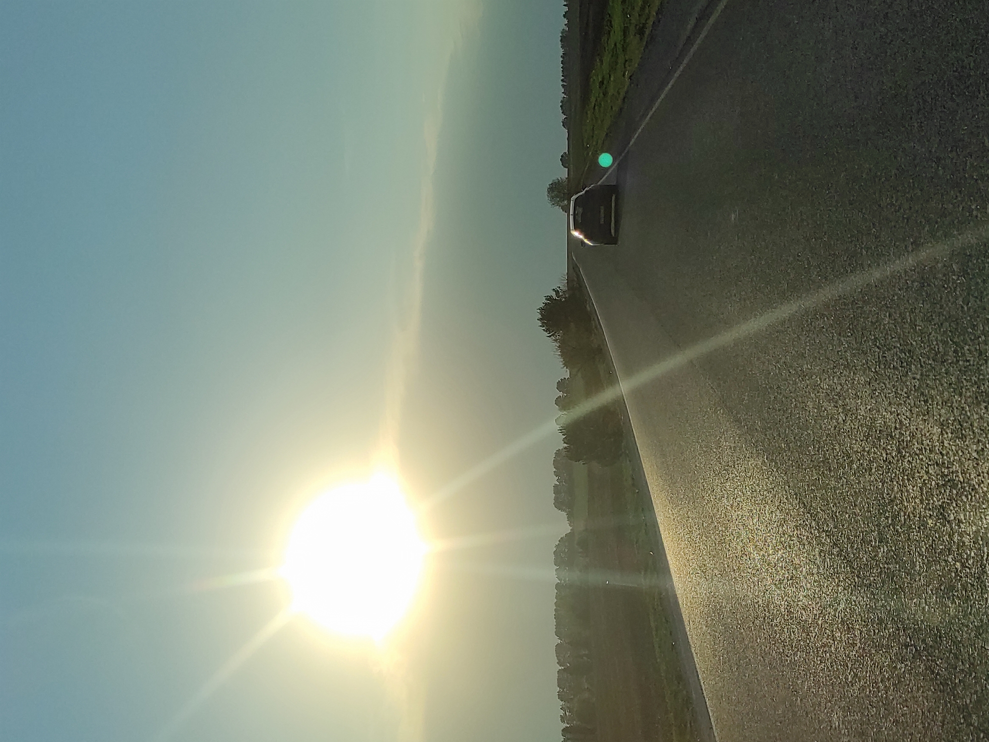 The image depicts a road with a car on it. The sun is bright in the sky, causing glare. The location is Перевозное, Russia. Trees line the side of the road.
