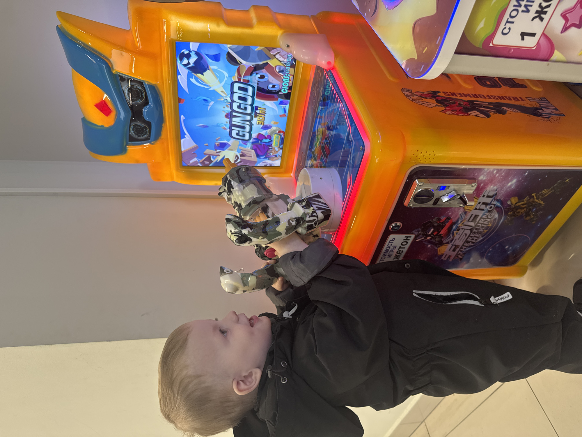 A young child, likely a toddler, is standing in front of a "GUN GOD" arcade game. The child is holding the light gun that is attached to the game. The arcade game is decorated with the colors yellow, blue, and orange. The game features graphics of the "TRANSFORMERS" franchise. The text on the game is in Russian, which may indicate the location of the image is in Saint Petersburg, Russia.