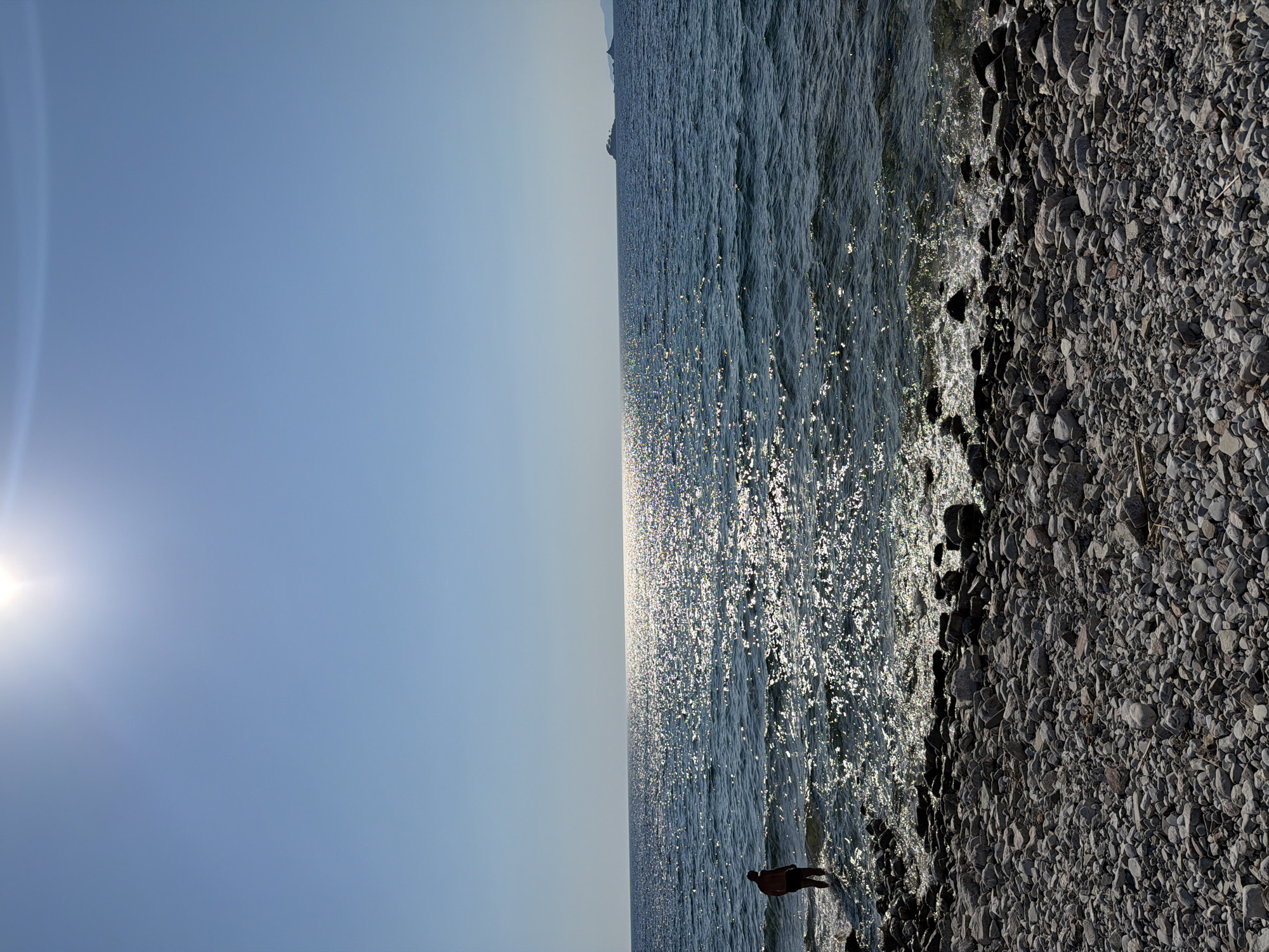 A person stands at the edge of a rocky shoreline, with the calm sea stretching out before them. The sun, visible in the upper left of the frame, casts a bright glare on the water, creating a shimmering effect. The sky is clear and blue, suggesting fair weather. The scene appears to be a tranquil beach, with the sun's rays indicating it's likely daytime, possibly late afternoon given the angle of the light. The water is a deep blue, with small waves gently lapping at the shore. The shoreline itself is composed of numerous gray and brown pebbles, varying in size. The person is silhouetted against the bright water, making their features indistinguishable. They appear to be looking out at the sea, perhaps contemplating the view or enjoying the peaceful atmosphere. There is no visible text in the image.