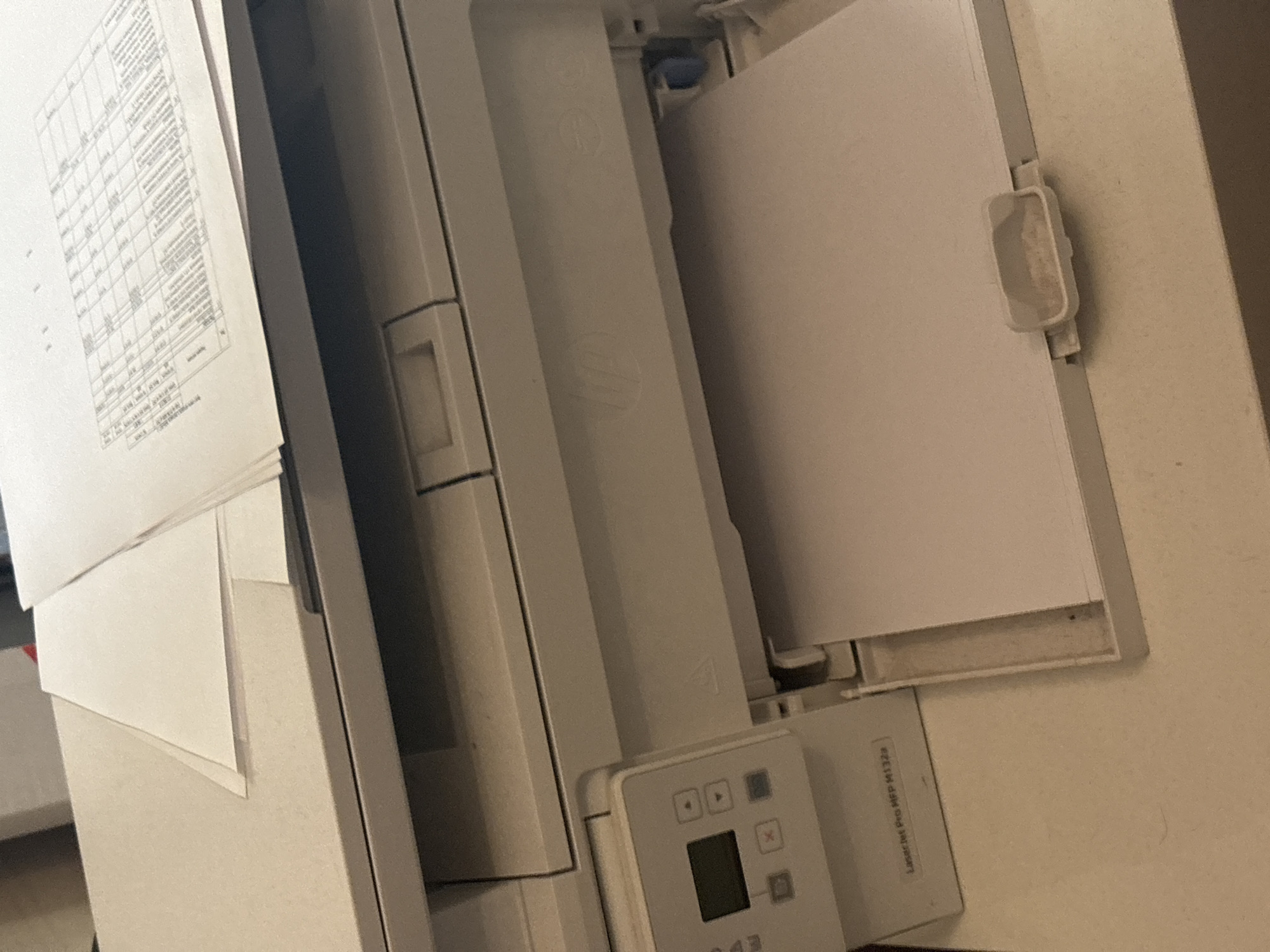The image shows a close-up view of a HP LaserJet Pro MFP M132a printer. The printer is located on a desk or table in what appears to be an office or home environment. Several sheets of paper are visible, with one sheet partially extended from the paper output tray. Another stack of papers, some with printed content that resembles a table or spreadsheet, is placed on top of the printer. The printer's control panel, with a small display screen and buttons, is visible on the left side of the machine. The overall lighting suggests an indoor setting. There is no information about the location in the city of Березка Снт (поварово), Russia within the image itself.