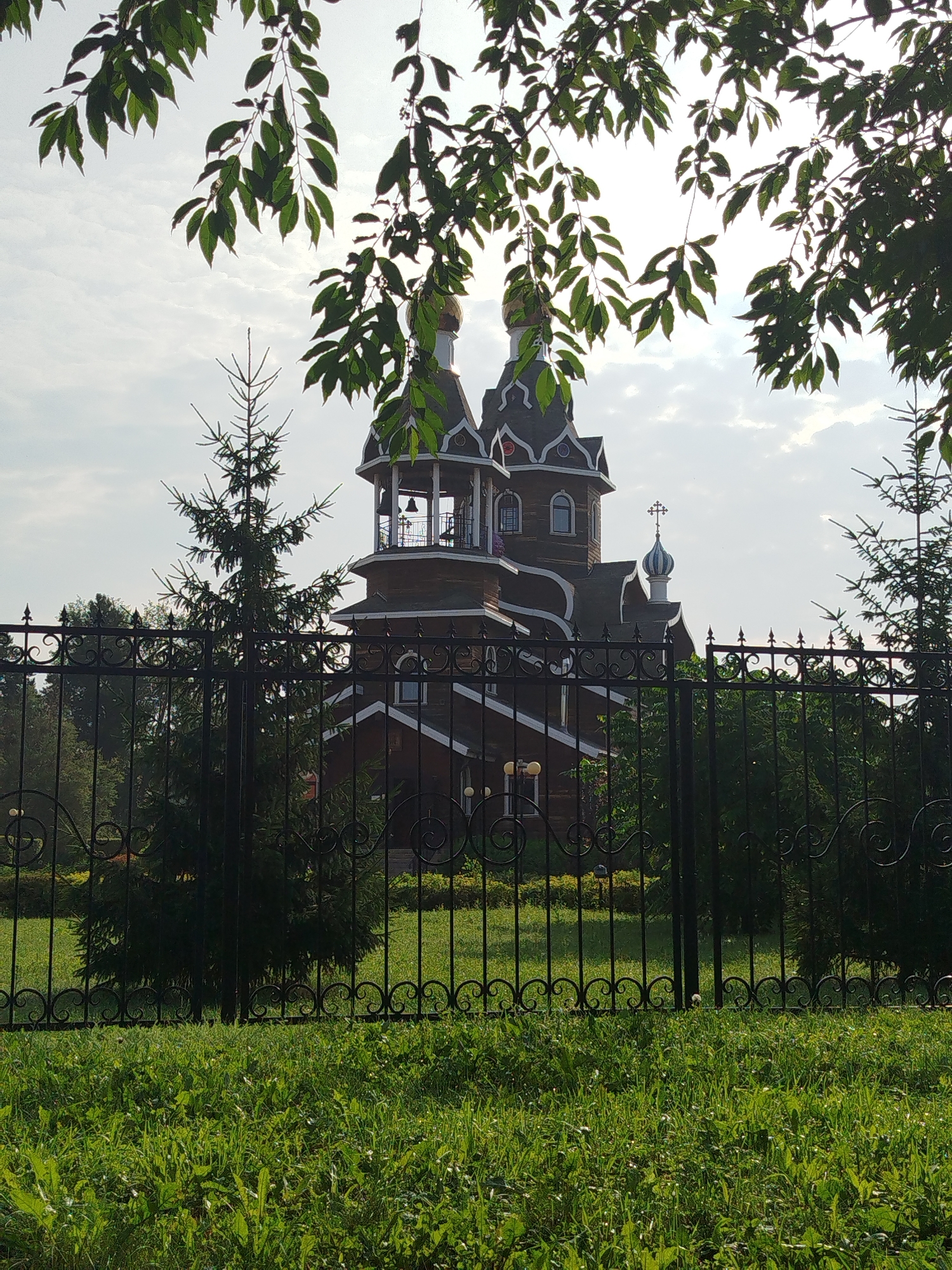 A sunlit, ornate wooden church with multiple onion domes and a bell tower is partially obscured by green foliage and a black metal fence. The church features dark wood construction with white trim and decorative elements. A cross adorns one of the domes. The foreground consists of lush green grass, and the background shows trees and a cloudy sky. The location is the Church of the Intercession of the Most Holy Mother of God in Berdsk, Russia.