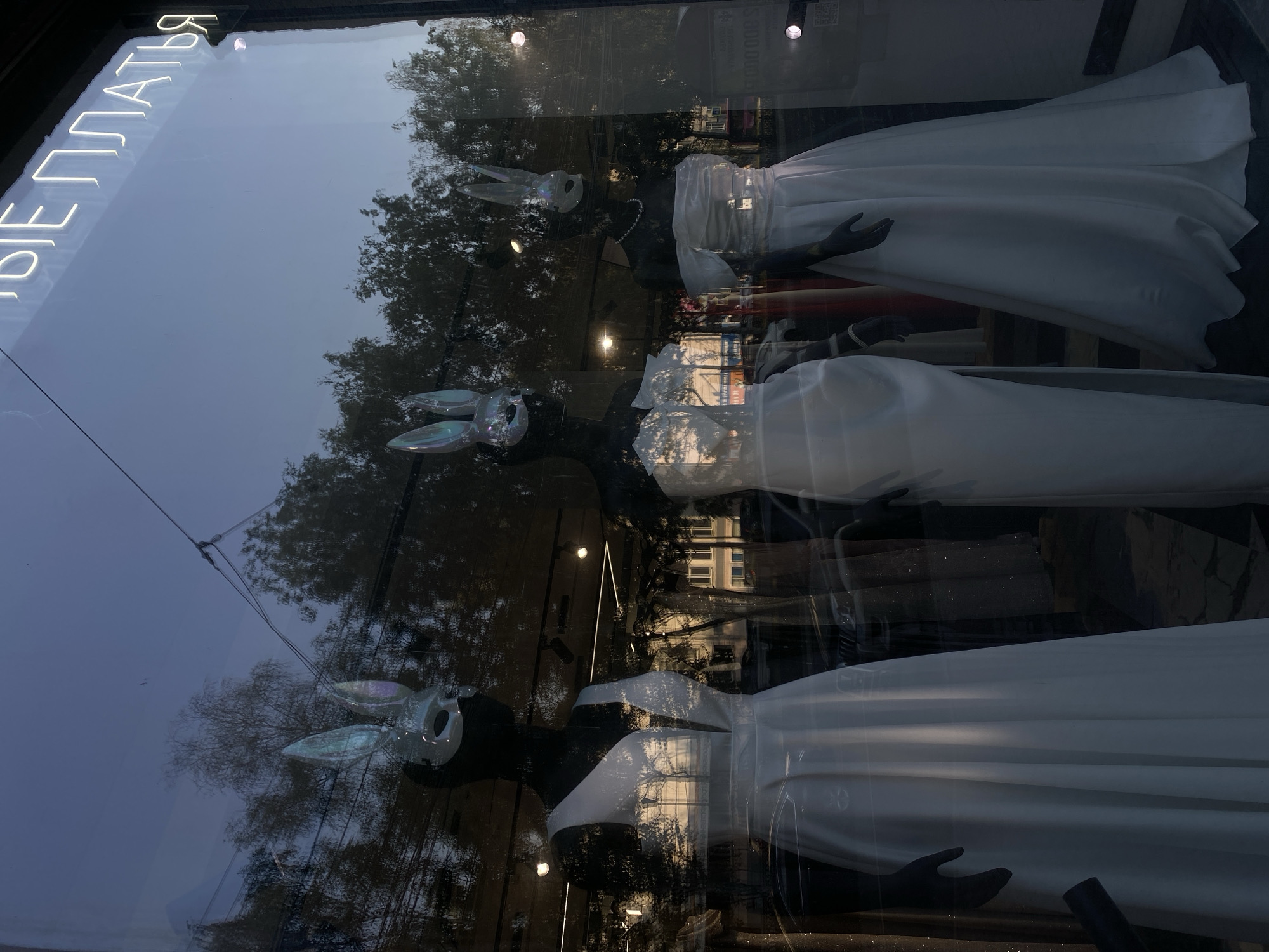 The image captures a storefront window display in Novosibirsk, Russia, showcasing three mannequins dressed in white gowns. Each mannequin wears a distinctive white mask with iridescent bunny ears. The leftmost mannequin is a full-body shot, displaying a sleeveless gown with a deep V-neck. The middle mannequin, also full-body, wears a strapless gown with draped fabric across the shoulders. The rightmost mannequin is partially visible, also in a strapless gown, adorned with a pearl necklace.
The window reflects the dusky sky, trees, and overhead wires, indicating an outdoor setting at dusk or early evening. The interior of the store, visible through the glass, contains additional clothing racks and lighting fixtures. A neon sign with Cyrillic lettering, which reads "НОВЫЕ ПЛАТЬЯ" (New Dresses), is prominently displayed at the top of the window. The overall scene suggests a boutique or dress shop.