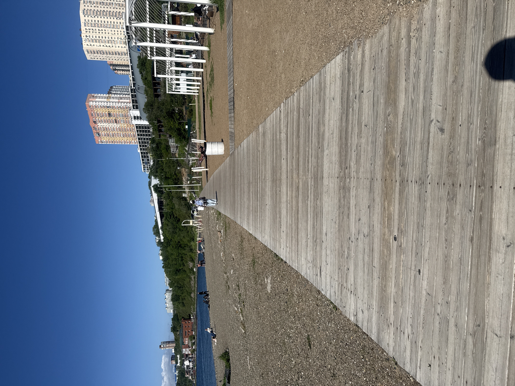 A wooden boardwalk extends into the distance on a sunny day at a beach in Vladivostok, Russia. To the left of the boardwalk is a gravelly beach with several people sitting and lying down. Beyond the beach is a body of water, and on the hills in the background are several apartment buildings and trees. To the right of the boardwalk is a sandy area with a white, open-air structure, and a trash can. A single person wearing a backpack walks away from the viewer on the boardwalk.