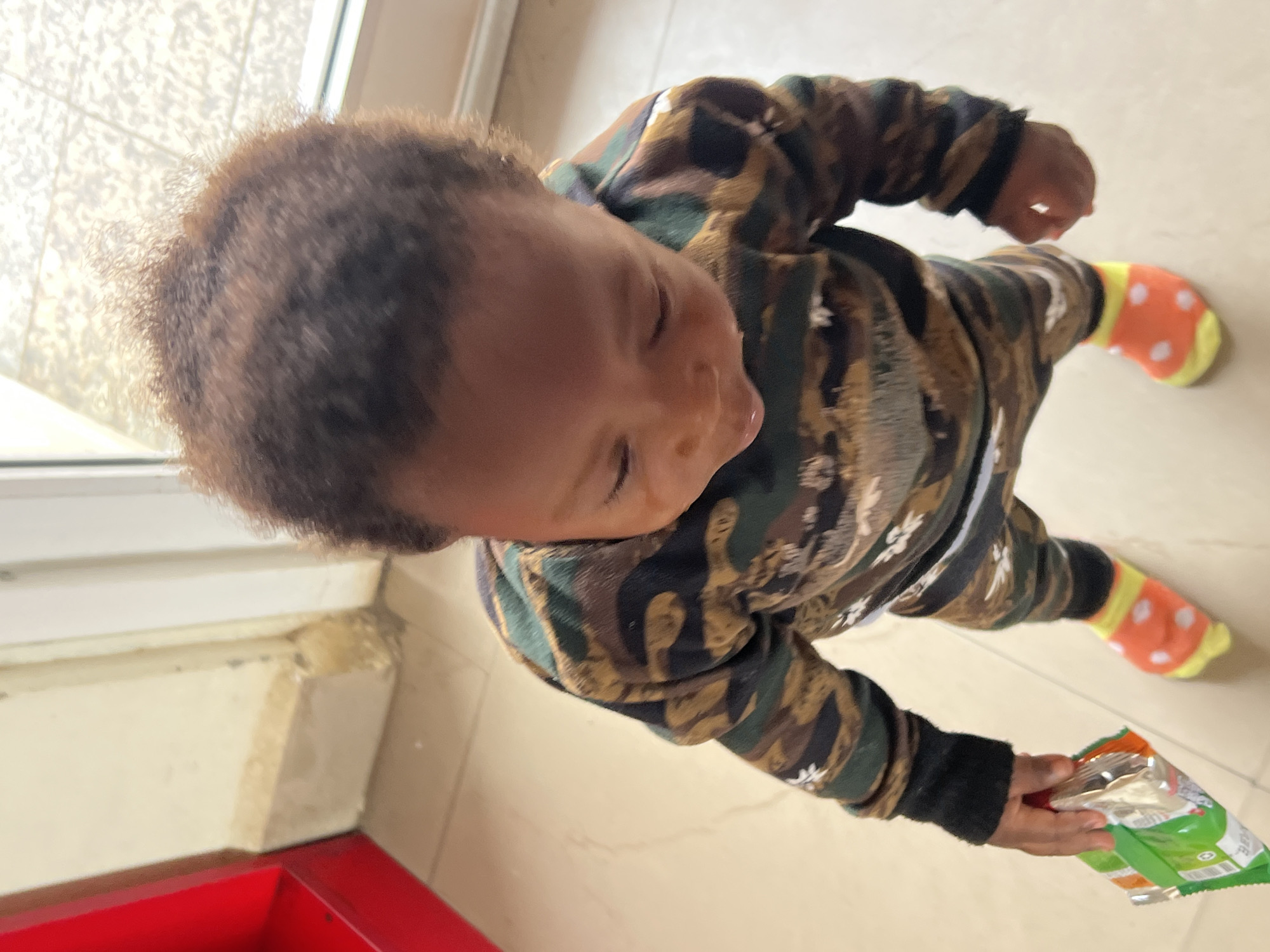 A young child is standing indoors on a light-colored tiled floor, viewed from directly above. The child is wearing a camouflage-patterned long-sleeved outfit and orange socks with white polka dots and yellow tops and heels. The child is holding a green and orange snack bag in their right hand. The child has dark, curly hair. A red and white window frame is visible in the upper left. It is not possible to identify the location in Gura Top, Nigeria from this image.