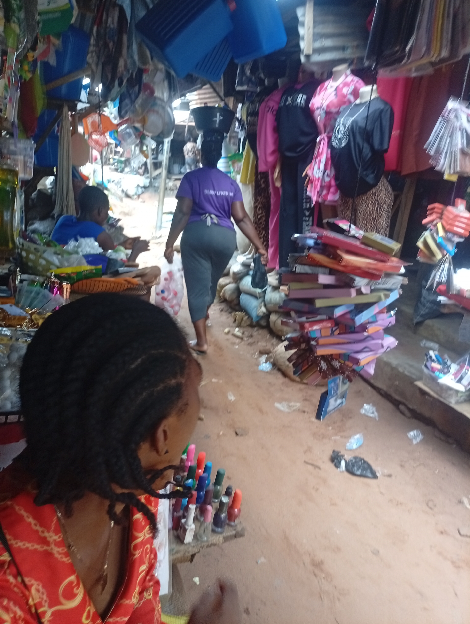 A woman in a purple t-shirt and grey pants walks away from the camera in a bustling outdoor market. She is carrying a clear plastic bag filled with items, possibly bottles. In the foreground, the back of another person's head is visible, with dark braided hair and a red patterned shirt. To the left, a child in a blue shirt sits in what appears to be a stall. The market is filled with various goods, including clothing on mannequins, boxes stacked high, and what looks like cosmetics or nail polish on a shelf. The ground is dirt. The scene appears to be during the daytime, with the overall atmosphere suggesting a lively and crowded commercial area. The t-shirt the woman is wearing has text that is partially visible, appearing to say "BUY / LIVES IN".