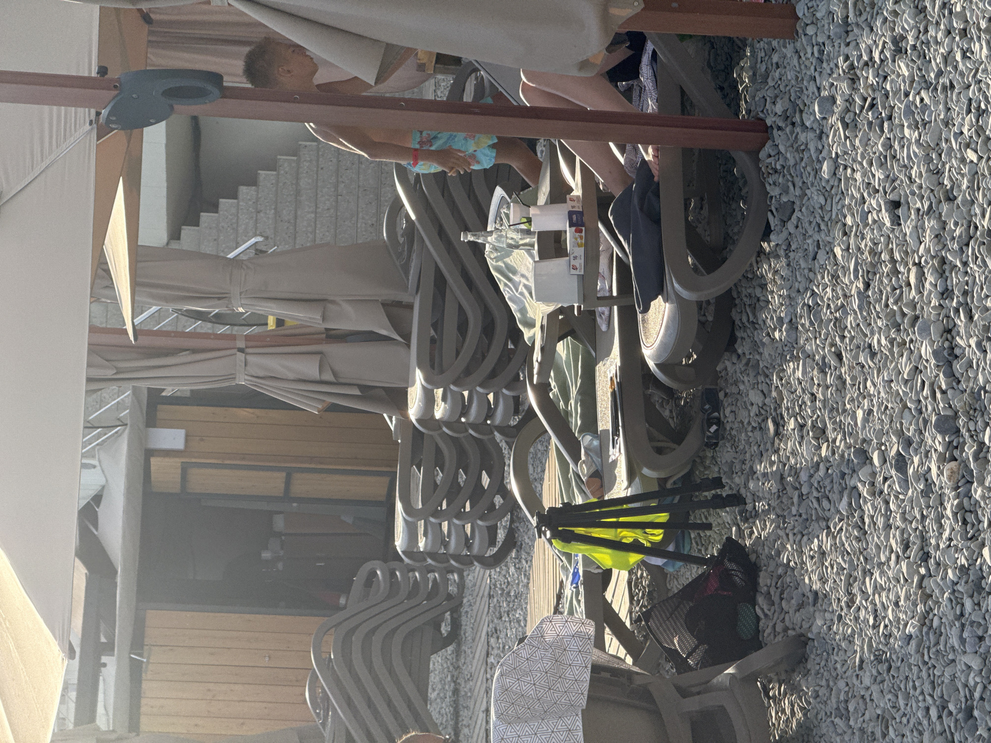 The image depicts a beach setting in Yalta, Ukraine. The foreground is dominated by a bed of light-colored pebbles, illuminated by the warm glow of the sun. In the mid-ground, a row of empty, stacked lounge chairs is visible, suggesting a public beach or resort area. These chairs are made of a light-colored material and are arranged on a wooden deck.
Behind the chairs, several people are partially visible. One individual, wearing blue patterned swim trunks, is seated and looking away. Another person's legs are visible, suggesting they are also seated. The people appear to be relaxed, enjoying a sunny day.
Notable details include the presence of umbrellas, some of which are open and providing shade, hinting at a warm and sunny day. A small table or cart is placed among the chairs, holding a few items that appear to be drinks and perhaps snacks. The overall atmosphere suggests a leisurely day at the beach. The background shows a concrete staircase leading upwards, possibly to a higher level of the resort or a building. The time of day appears to be late afternoon, given the angle of the sunlight casting long shadows. There is no visible text in the image.