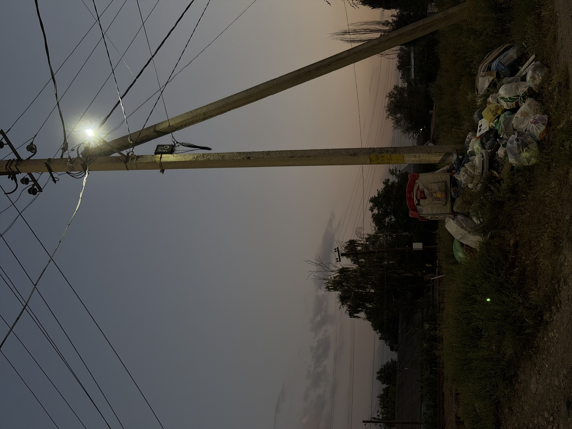 This media file depicts an outdoor scene at dusk, with a low-lying, overcast sky in muted tones of gray and faint orange. The primary subjects are utility poles with numerous electrical wires crisscrossing the frame. One pole is illuminated by a bright, focused light.
To the right of the utility poles, a large pile of trash is visible, consisting of various plastic bags, containers, and discarded items. A small, red and white object, possibly a child's toy or a piece of furniture, is nestled within the garbage. The ground is covered in dry grass and dirt.
In the background, silhouetted trees and vegetation are discernible. The overall impression is one of neglect and disarray, juxtaposed with the mundane infrastructure of power lines. The time of day is clearly evening, as indicated by the dim, diffused light and the dusky sky. There are no visible people in the image.
There is some yellow text on a white background visible on one of the utility poles, but it is too blurry to be read. Additionally, there are some black markings on a rectangular object attached to one of the poles, also unreadable.
The scene appears to be in a semi-urban or rural setting, possibly on the outskirts of a populated area, given the presence of both infrastructure and accumulated waste. The weather appears to be clear but with heavy cloud cover contributing to the dim light. No specific emotion is conveyed beyond the somber mood suggested by the twilight and the waste.