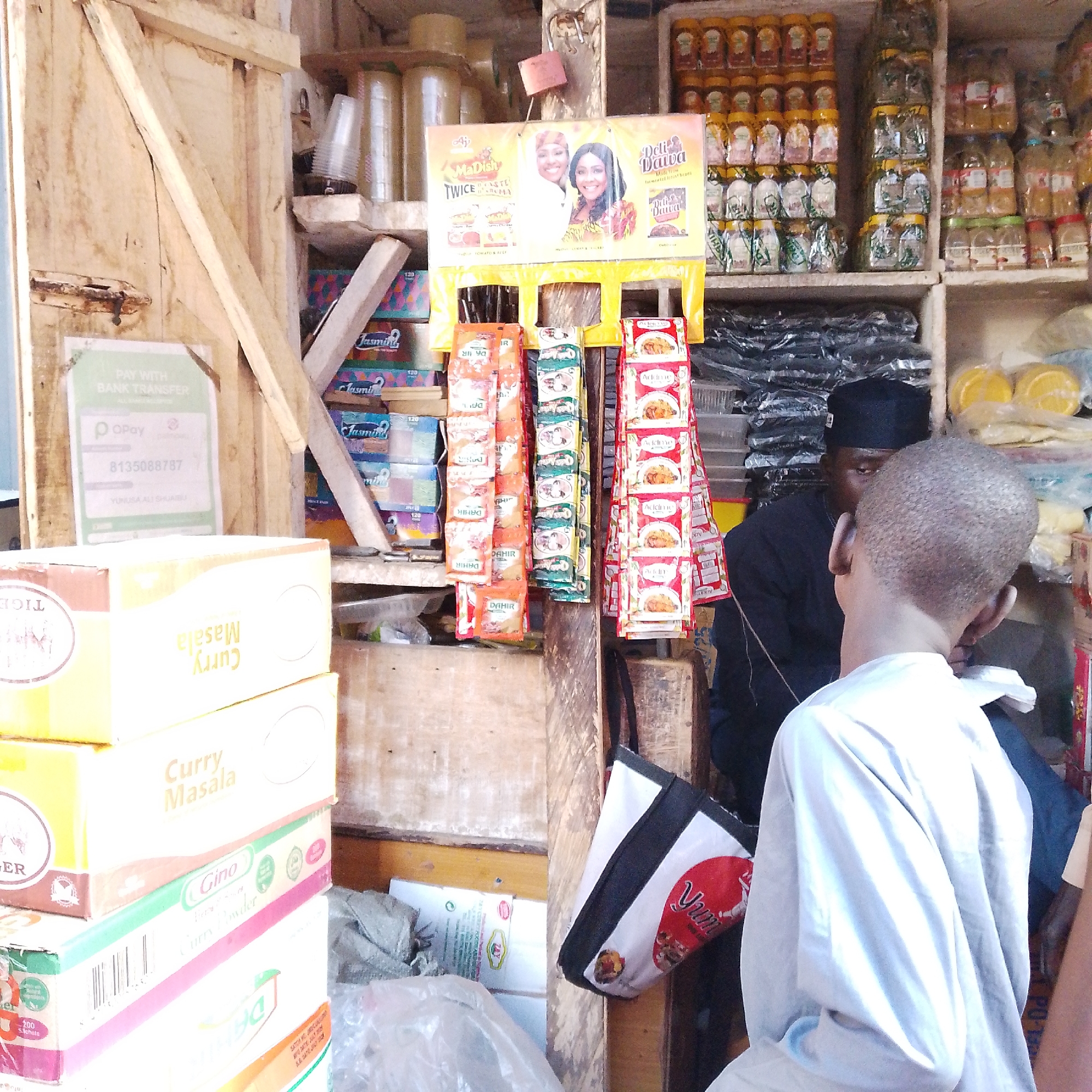 The image depicts a small shop in Arawa, Nigeria, likely a food stall or a convenience store. In the foreground, a young boy is seen from the back, wearing a light-colored garment. He is facing away from the camera, towards the shop's interior. To his left, stacked boxes are visible, with one prominently displaying "Curry Masala" in yellow text. Above these boxes, a colorful advertisement featuring two women promoting "MaDish TWICE THE TASTE TWICE THE AROMA" is displayed.
The shop's interior is filled with various goods. Shelves are stocked with jars of what appear to be spices or condiments, some with yellow lids, others with clear lids and white labels. Rolls of tape are stacked on a wooden structure. Several packets of seasoning or spices are hanging from a central pole. These packets have colorful designs and text.
A man wearing a black cap and a dark garment is partially visible behind the young boy, seemingly attending to customers or organizing goods. The shop is constructed with rough wooden planks, giving it a rustic appearance.
To the left of the scene, a sign reads "PAY WITH BANK TRANSFER" and displays an OPay logo with a phone number. Other miscellaneous items, including plastic bags and what seem to be packaged food items, are scattered throughout the shop, indicating a bustling marketplace environment. The overall scene suggests a typical local Nigerian retail setting.