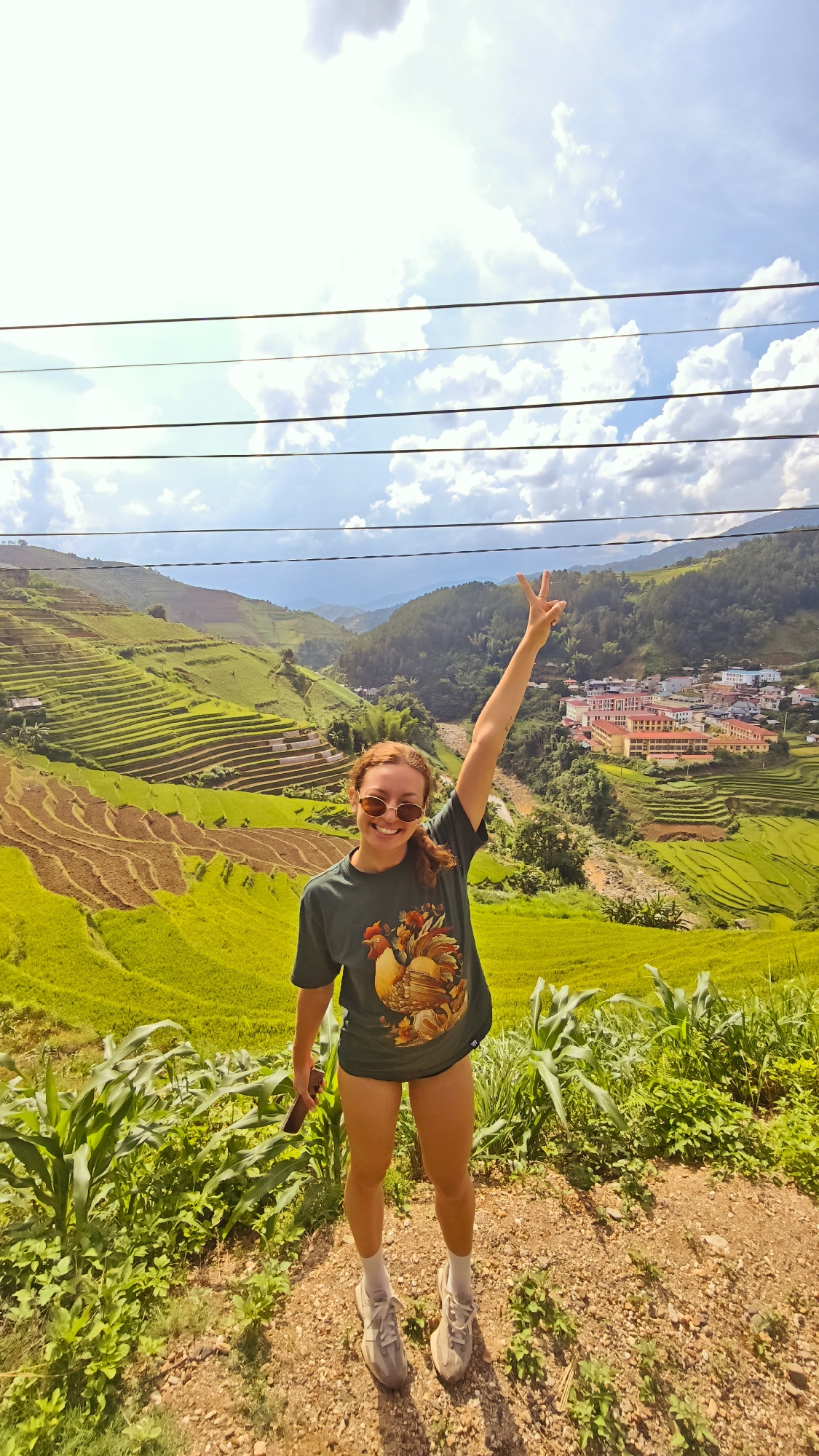 This full-length portrait captures a smiling young woman posing joyfully against a vast and scenic landscape in Lao Chai, Vietnam. She has reddish-brown hair tied in a low ponytail, wears round sunglasses, a dark green t-shirt with a colorful rooster graphic, dark shorts, white socks, and light grey athletic shoes. Her right arm is raised, forming a "peace" sign, while she holds a smartphone in her left hand. Her wide smile indicates happiness and enjoyment of the moment.
The setting is characterized by extensive terraced rice fields that dominate the mid-ground, showcasing both vibrant green paddies and patches of cultivated brown soil. A valley runs through the landscape, beyond which a village with buildings featuring orange-red roofs and lighter walls is visible. Rolling green mountains rise in the distance under a bright blue sky with scattered white clouds. Several dark power lines stretch horizontally across the upper part of the image. The foreground features dry, dusty ground with some sparse green vegetation, including what appear to be corn stalks.
The activity depicted is a person posing for a photograph, likely during travel or a hike, enjoying the stunning agricultural and mountainous scenery. The time of day appears to be bright daylight, possibly late morning or early afternoon, under sunny and clear weather conditions. The terraced fields are a strong visual cue for the region. The colorful rooster on her shirt is a notable detail, but no explicit text is visible.