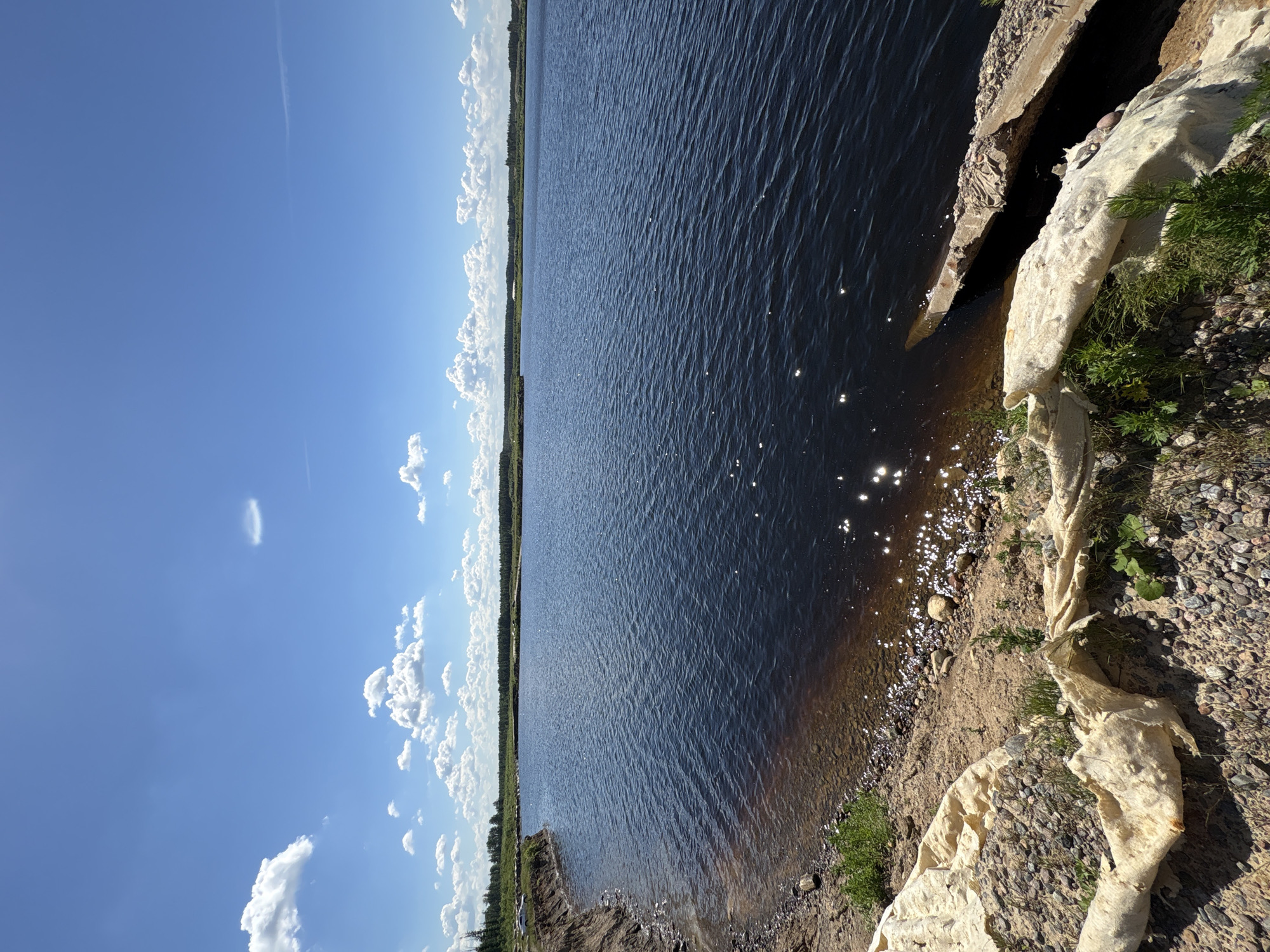 The image depicts a waterfront scene in Yoksolovo, Russia, under a clear blue sky with scattered white clouds. The foreground shows a rocky shoreline with some scattered debris, including what appears to be a large piece of torn, beige fabric or tarp. Further down the shore, there is a pile of rocks and some green vegetation. The dark blue water of a lake or river occupies the majority of the frame, with gentle ripples reflecting the sunlight. In the distance, a line of trees forms the horizon, with more trees and greenery visible on the far bank. The overall environment suggests an outdoor, natural setting. There are no people or animals visible in the image. The context of the location being in Yoksolovo, Russia, provides a geographical identifier for the scene.