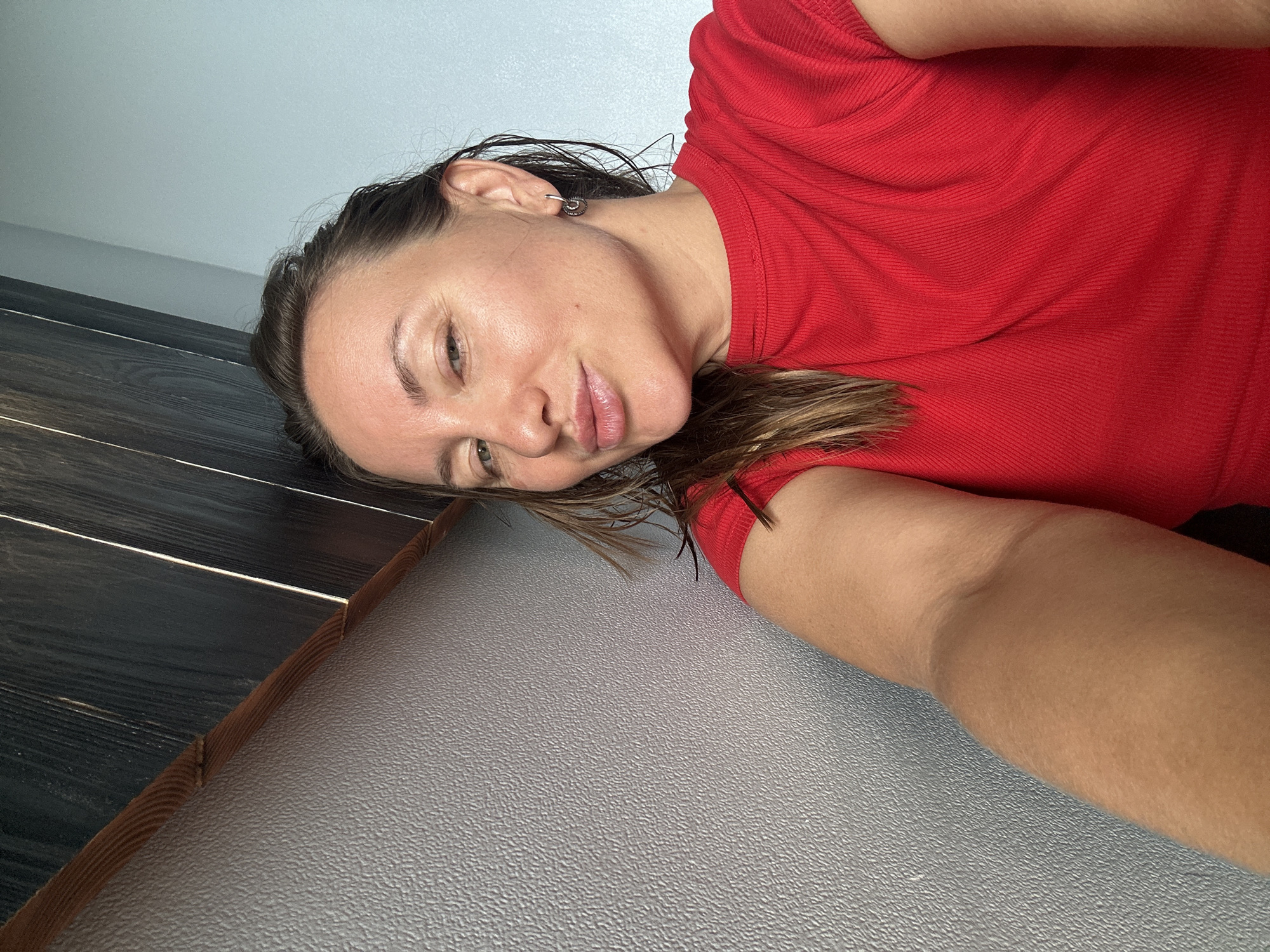 The image is a selfie of a woman in her early 30s. She has light skin, brown hair, and is wearing a red t-shirt. She is looking at the camera with her lips pursed. The background is a wall with a textured gray finish and a wooden panel above it. The scene appears to be indoors.