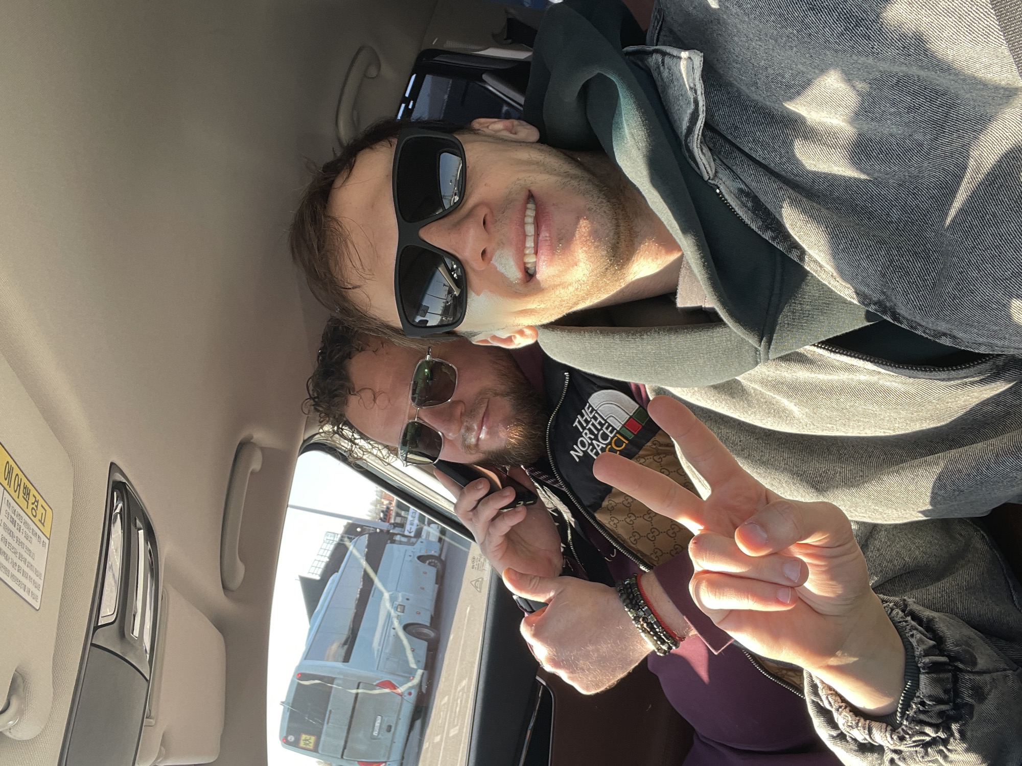 Two men are in a car. The man closest to the camera is on a phone call, giving a peace sign with his right hand, and wearing sunglasses, a North Face vest over a purple shirt, and a bracelet. The man next to him is smiling, wearing sunglasses and a gray jacket, with white cream applied to his nose. Outside the car, a white bus is visible. The scene appears to be Kudrovo, Russia.