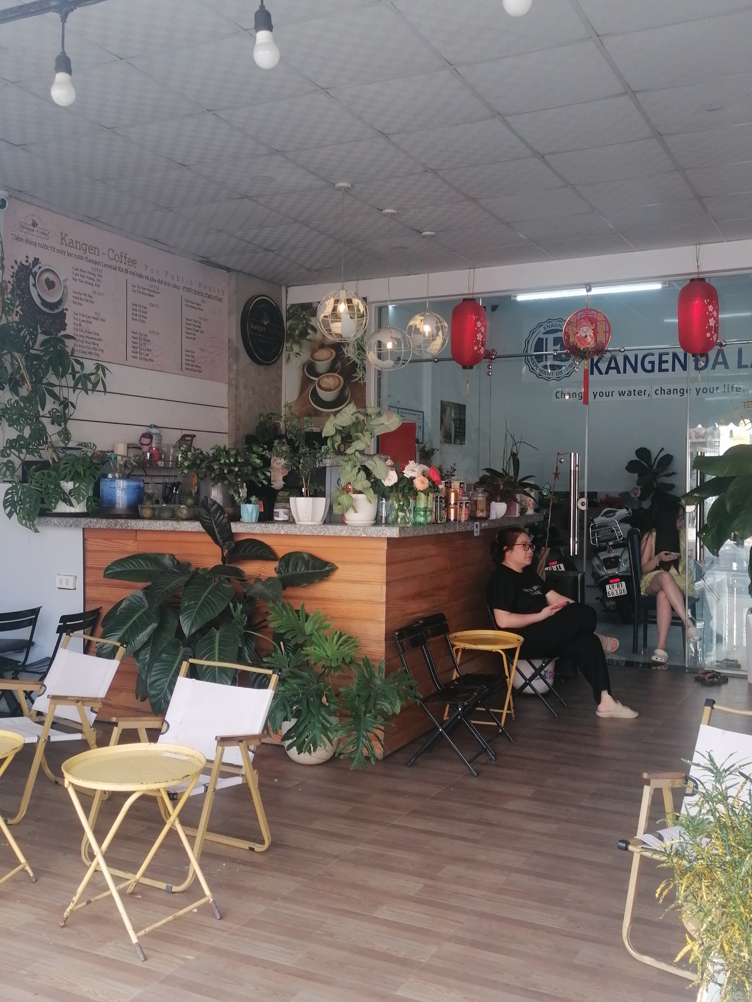 The image shows the interior of a Kangen Coffee shop in Da Lat, Vietnam. A menu board displays coffee and tea options in Vietnamese. There is a wooden counter with various plants. Customers are seated at tables, one wearing glasses. Red lanterns and globe-shaped lights hang from the ceiling. A motorcycle is visible through the glass entrance. The shop's name, "KANGEN DA LA," is displayed on the glass, along with "Change your water, change your life."