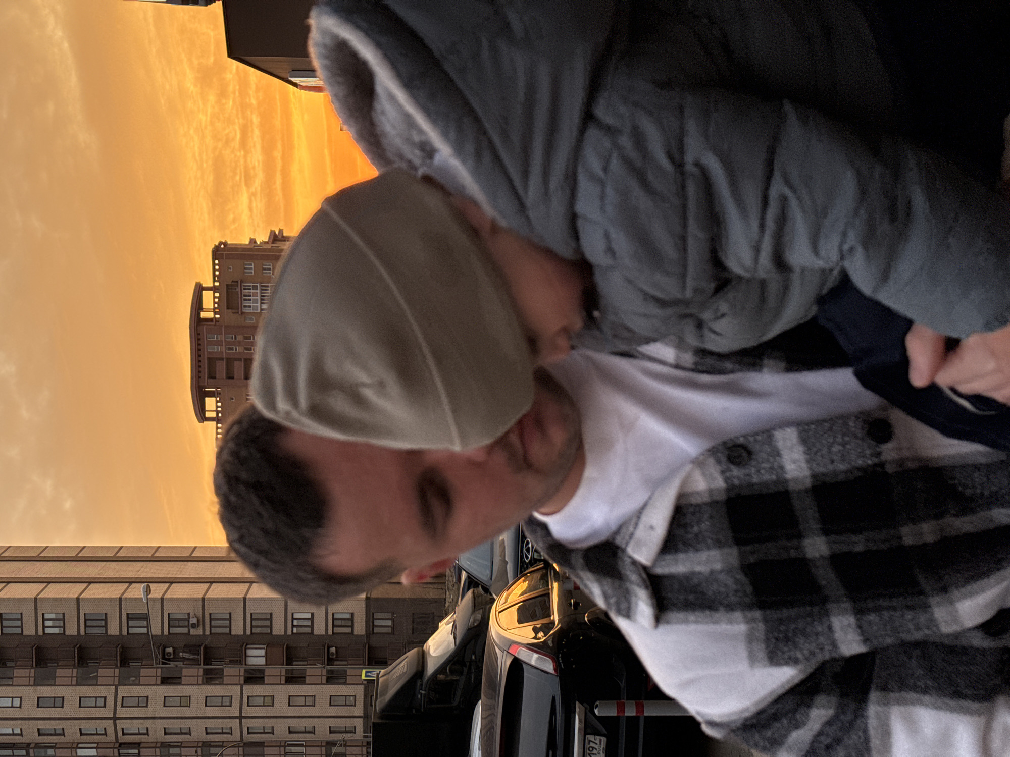 The image captures an adult male holding a young child outdoors during sunset in Moscow, Russia. The man, positioned centrally, is seen from the chest up, with short dark hair, wearing a white t-shirt and a black-and-white plaid long-sleeved shirt. He looks downwards, seemingly at the child. The child, held in the man's left arm, wears a light brown beanie and a grey puffer jacket with a fur-lined hood, and is also looking downwards, obscuring their face.
The background consists of an urban setting illuminated by the warm, orange and yellow hues of a sunset sky. To the left, a tall, multi-story residential building constructed with brown brick and numerous windows and balconies is visible. Further back, another lighter-colored building with a distinctive rooftop structure is present. Parked vehicles are visible behind the subjects, including a black commercial van and a dark-colored Ford Focus hatchback. A partially visible Russian license plate on the Ford Focus reads "197 RUS," identifying the location as Moscow. Red and white striped bollards are seen on the pavement next to the parked cars. A street lamp and a pedestrian crossing sign on a pole are also discernible near the tall building.