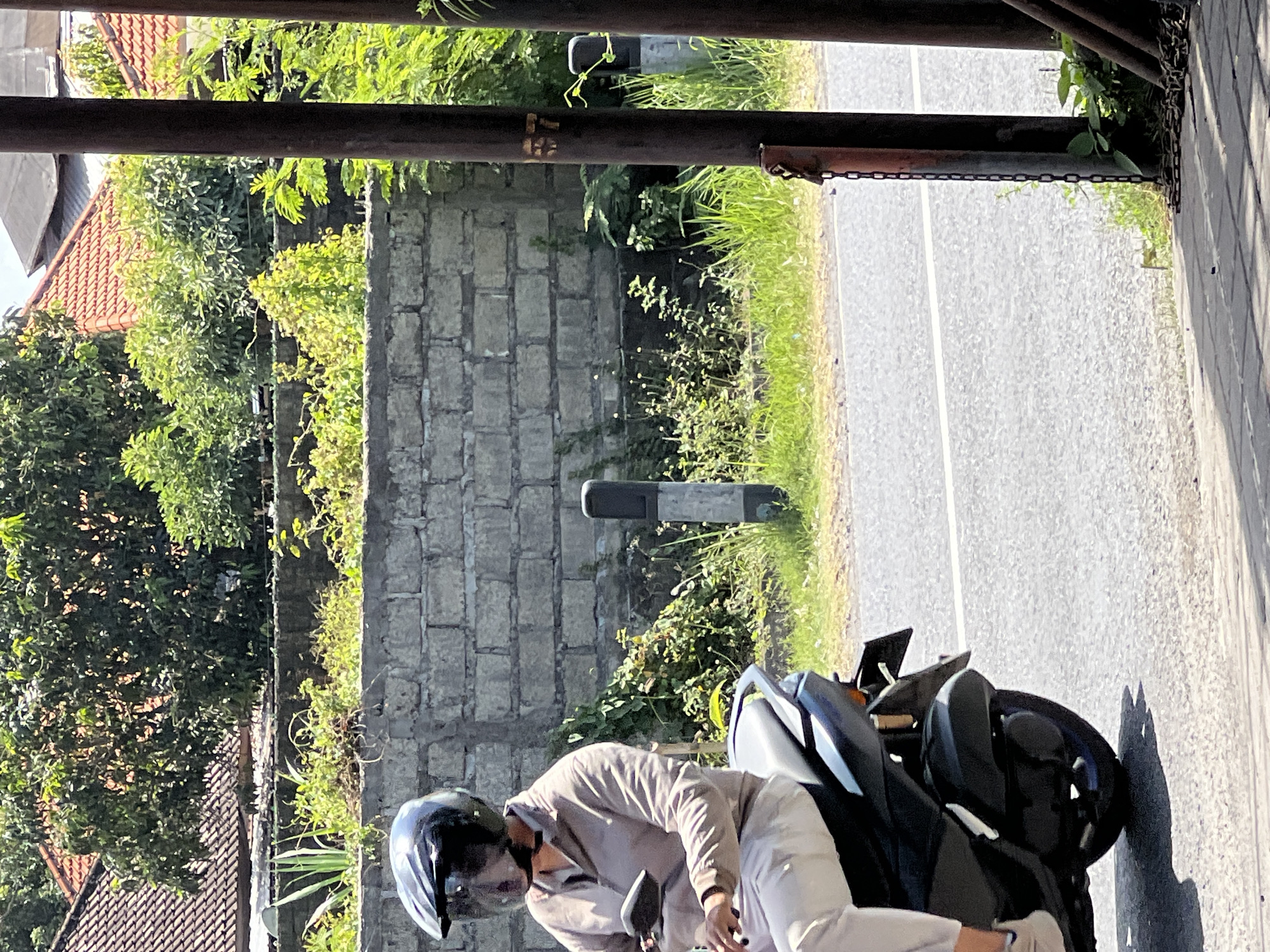 A person wearing a black helmet with a clear visor is riding a dark-colored motorcycle on a paved road. The person is wearing a light-colored long-sleeved shirt and light-colored pants. The motorcycle has a black seat and a visible rear wheel. In the background, there is a low wall constructed of grey bricks, overgrown with green vegetation. Beyond the wall, there are trees and buildings with red tile roofs. Two dark, cylindrical posts stand in the foreground on the right side of the image, with a rusty chain hanging from one of them. The scene appears to be in Denpasar, Indonesia, characterized by the architecture and lush vegetation.