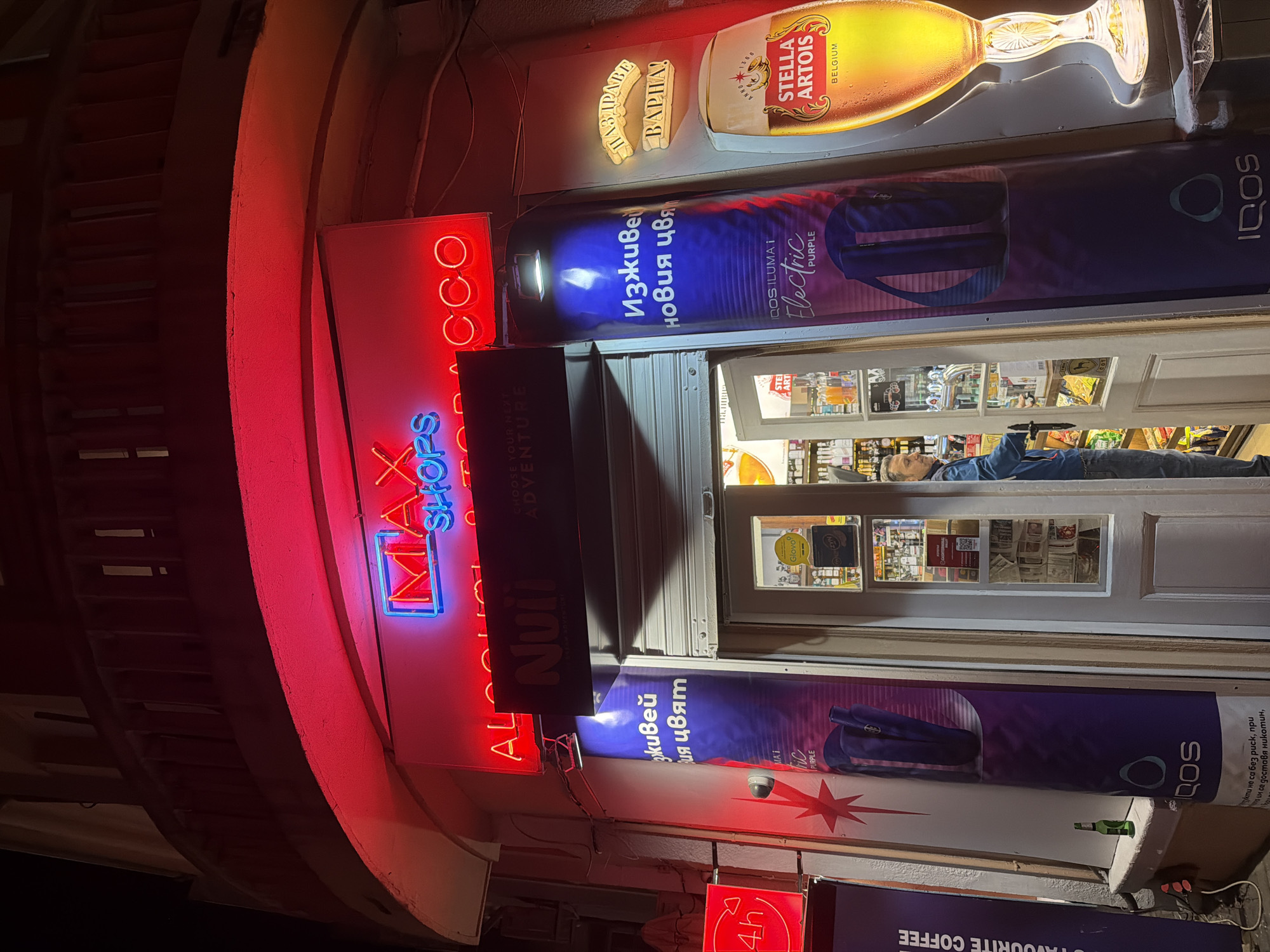 The image shows the exterior of a shop in Varna, Bulgaria, at night. The shop is identifiable by its neon signs that read "MAX SHOPS ALCO TOCCO Nui", as well as advertisements for Stella Artois beer and IQOS electronic cigarettes. A person is visible inside the shop, near the doorway, wearing a blue jacket and jeans. The shop window displays various items for sale. A neon sign reading "4h" indicates opening hours. The architecture includes a curved wall to the left of the shop's entrance.
