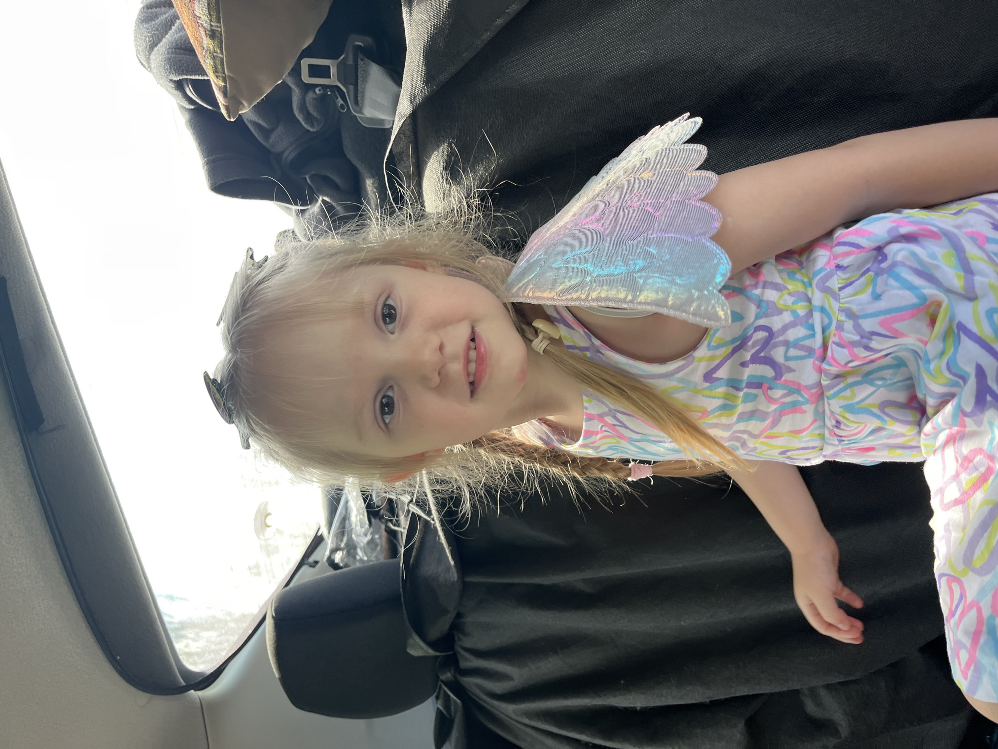 A young girl with blonde hair in pigtails and a colorful dress with iridescent fairy wings is sitting in the back of a car. She is looking at the camera with a slight smile. The car's interior is visible, with black seats and a bright window behind the girl. The image is taken from a medium shot, focusing on the girl's face and upper body. There is no visible indication that this image was taken in Kovriga, Russia.