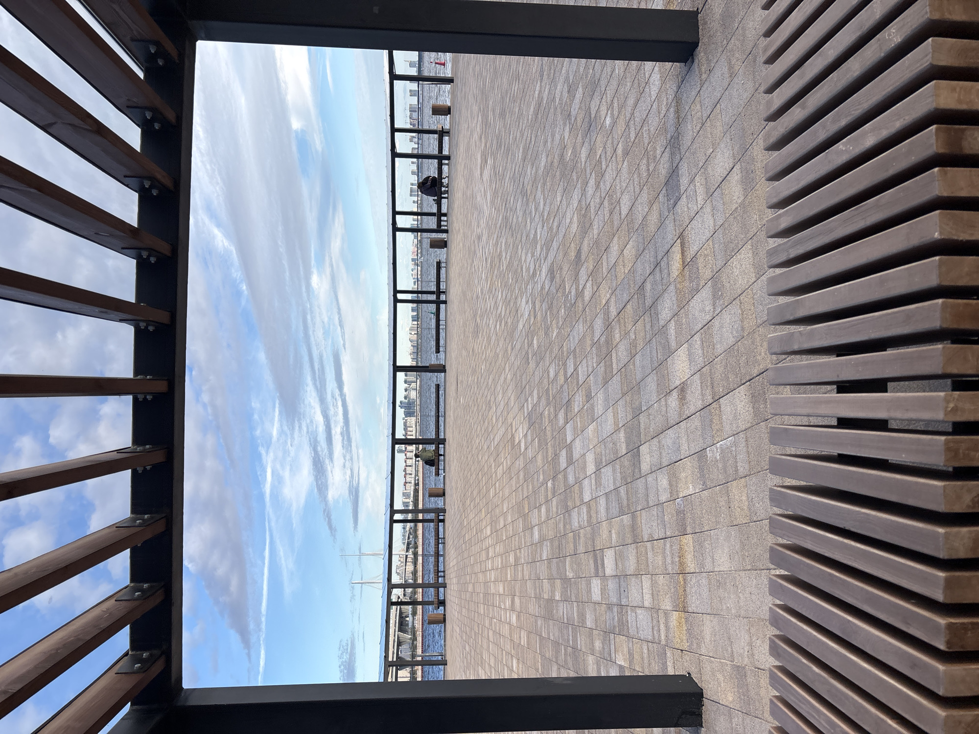 The image shows a public waterfront plaza in Saint Petersburg, Russia, viewed through the frame of a wooden pergola. The plaza is paved with light-colored square tiles. In the middle ground, there is a body of water, likely the Gulf of Finland, with a bridge and a cityscape in the distance. Along the waterfront, there are several benches and trash receptacles. Two individuals are seated on a bench, facing the water. The sky is partly cloudy with a mix of blue and white. In the foreground, a portion of a wooden bench is visible, with its slats running horizontally across the bottom of the frame.