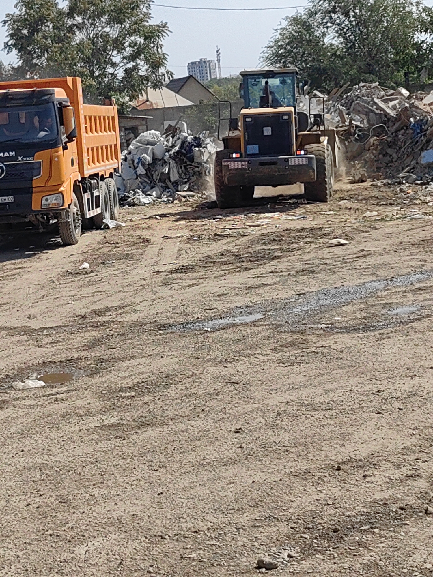 An orange dump truck is parked on a dirt road next to a pile of debris. To the right of the dump truck, a yellow front-end loader is in motion, with dust kicked up around its wheels and bucket. The loader appears to be pushing or moving the debris. In the background, there are trees and buildings, suggesting an urban or semi-urban setting. The sky is visible and appears clear. The overall scene depicts a construction or demolition site, likely in Shymkent, Kazakhstan, given the context.