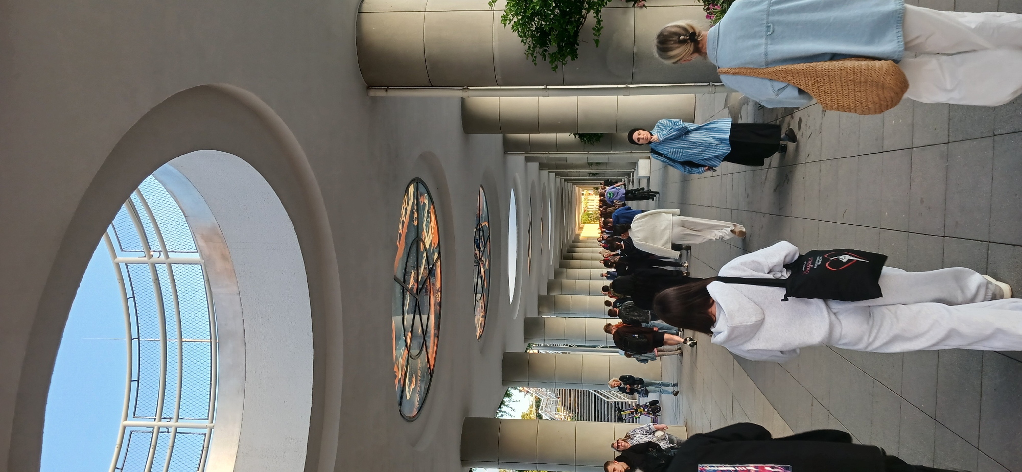 The image depicts a wide, covered pedestrian walkway with a series of cylindrical concrete columns supporting an arched ceiling. Several circular stained-glass panels are embedded in the ceiling. The walkway is populated by a diverse group of people, many of whom are walking in both directions. The ground is paved with large, light-colored rectangular tiles. To the left, a staircase leads upwards, with a white railing and mesh fencing. The environment appears to be outdoors, under a clear sky, suggesting a public space within a city. The presence of a bicycle and what appears to be an electric scooter suggests a place where such transportation is common. The overall atmosphere is one of casual pedestrian activity.