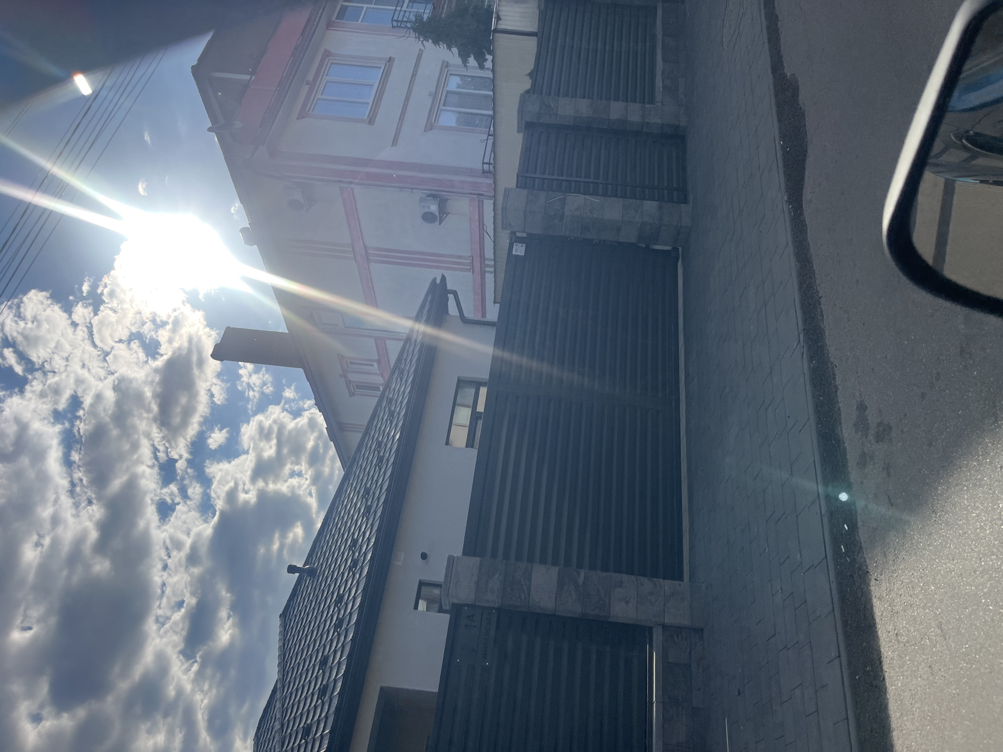 In the city of Storozhnytsia, Ukraine, a two-story building with a light-colored facade and pink accents is visible under a bright, partly cloudy sky. The sun's rays create a lens flare effect, obscuring some details of the upper portion of the building. A dark, ribbed metal gate runs horizontally across the foreground, supported by stone pillars. To the left, a portion of another building with a dark, shingled roof is visible, also behind a similar gate. The street in front of the gate is paved with gray interlocking stones. A sidewalk runs along the street, and a dark asphalt road is also present. The scene appears to be captured from a vehicle, as a car's side mirror is visible in the lower right corner of the frame, reflecting part of the car.