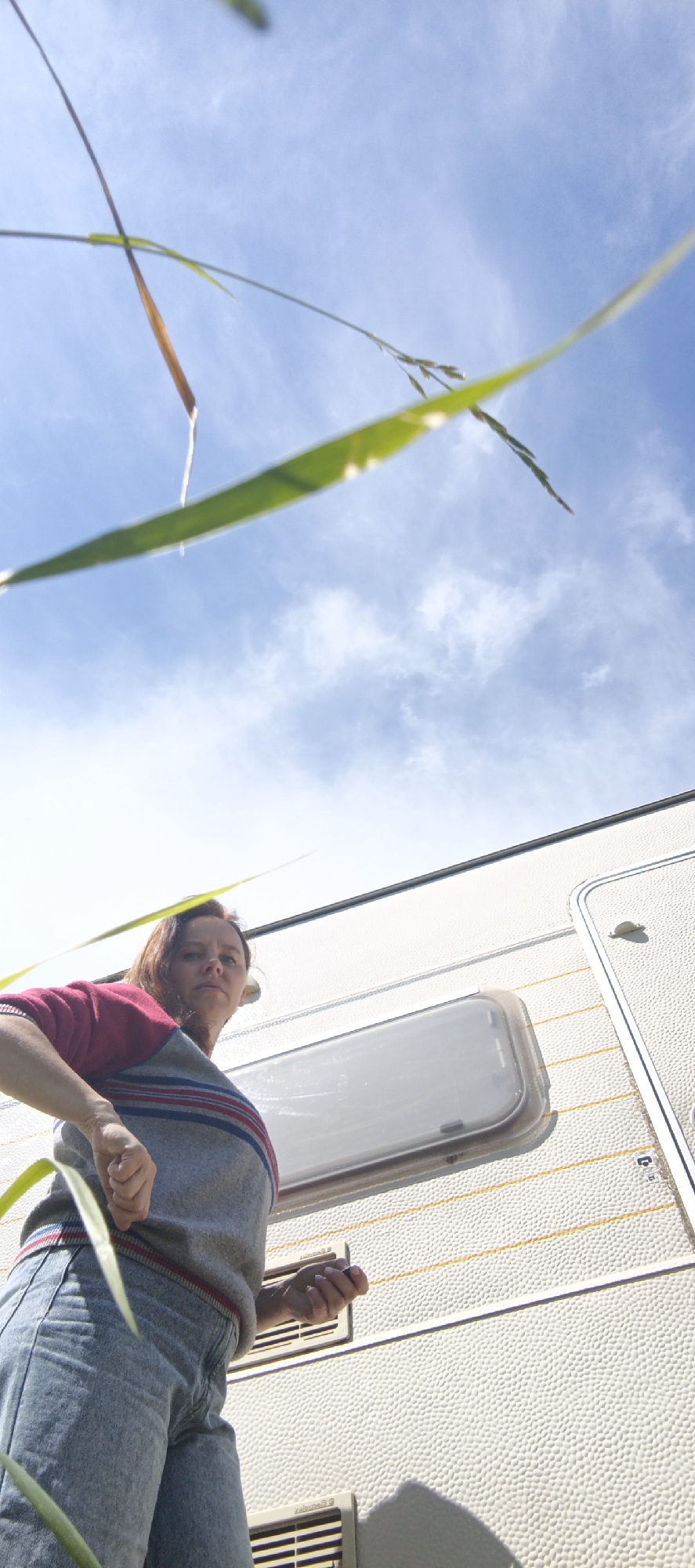A woman stands in front of a white caravan, viewed from a low angle looking up. She has auburn hair and is wearing a grey sweater with red and blue stripes and blue jeans. The caravan has a rectangular window and ventilation grates. The background is a bright blue sky with scattered white clouds, and blades of grass are in the foreground, partially obscuring the view. The image appears to be taken outdoors during daylight. There is no visible indication of the specific location within Chaikovsky, Russia.