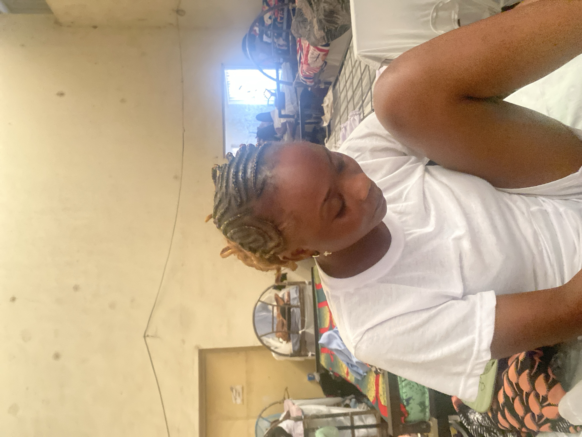 A young Black woman with braided hair and a white t-shirt is seated in a room. She is looking down and has her right leg bent up towards her chest. The room appears to be a dormitory or shared living space, with multiple beds visible in the background. The beds are made with colorful linens and some have mosquito nets. The walls are a pale yellow and show signs of wear and tear. There is a window in the background that lets in natural light. The overall impression is one of a simple and functional living environment. The image was taken in Jimeta, Nigeria.