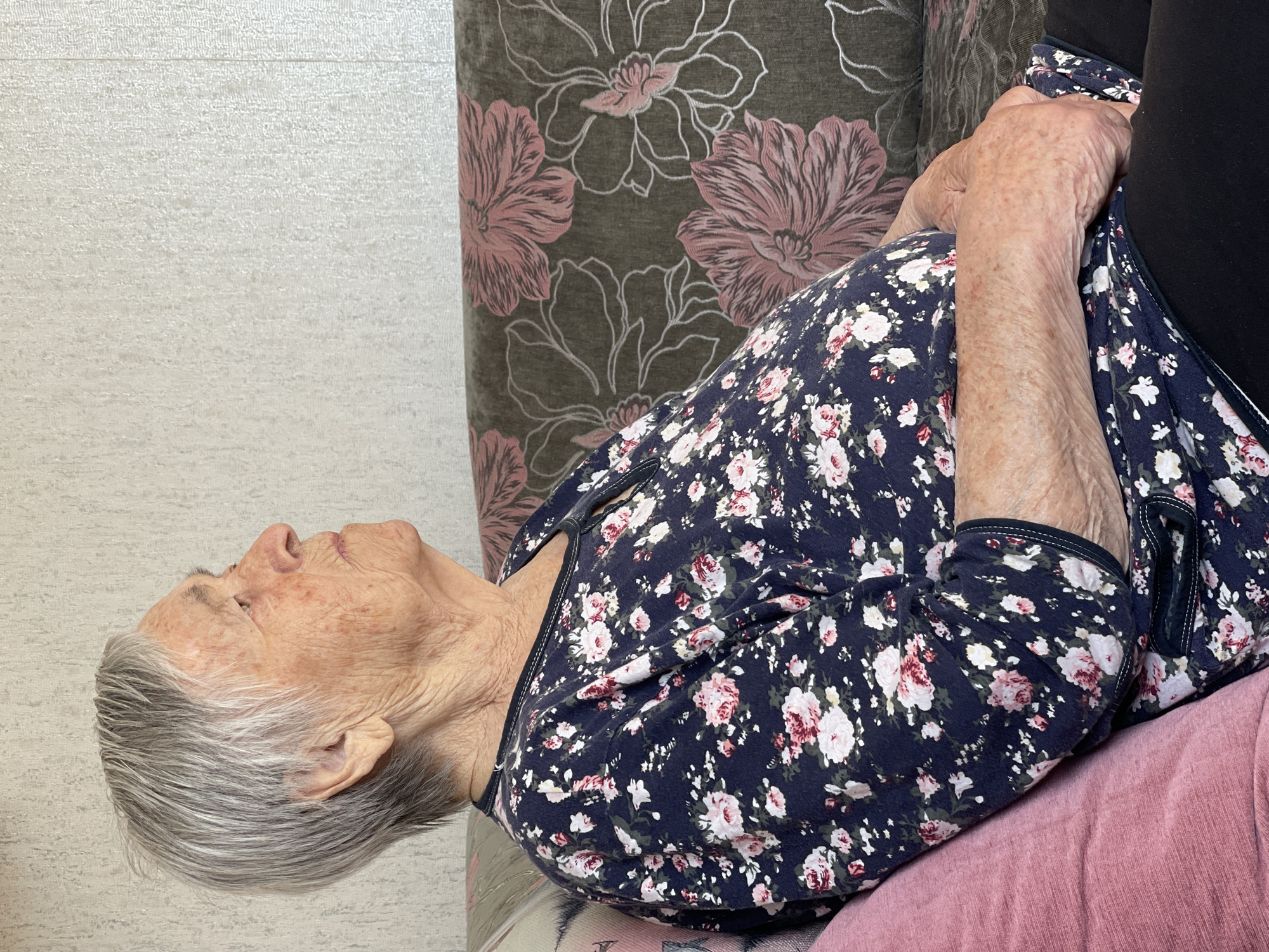 An elderly woman with gray hair is reclining on a cushioned surface with a floral pattern, appearing to be a sofa or chair. She is wearing a dark blue floral print top. Her hands are clasped together. She is situated next to a wall with a light-colored, textured surface. In the background, a patterned cushion with large pink floral designs is visible. The location of the image is Перевозное, Russia.