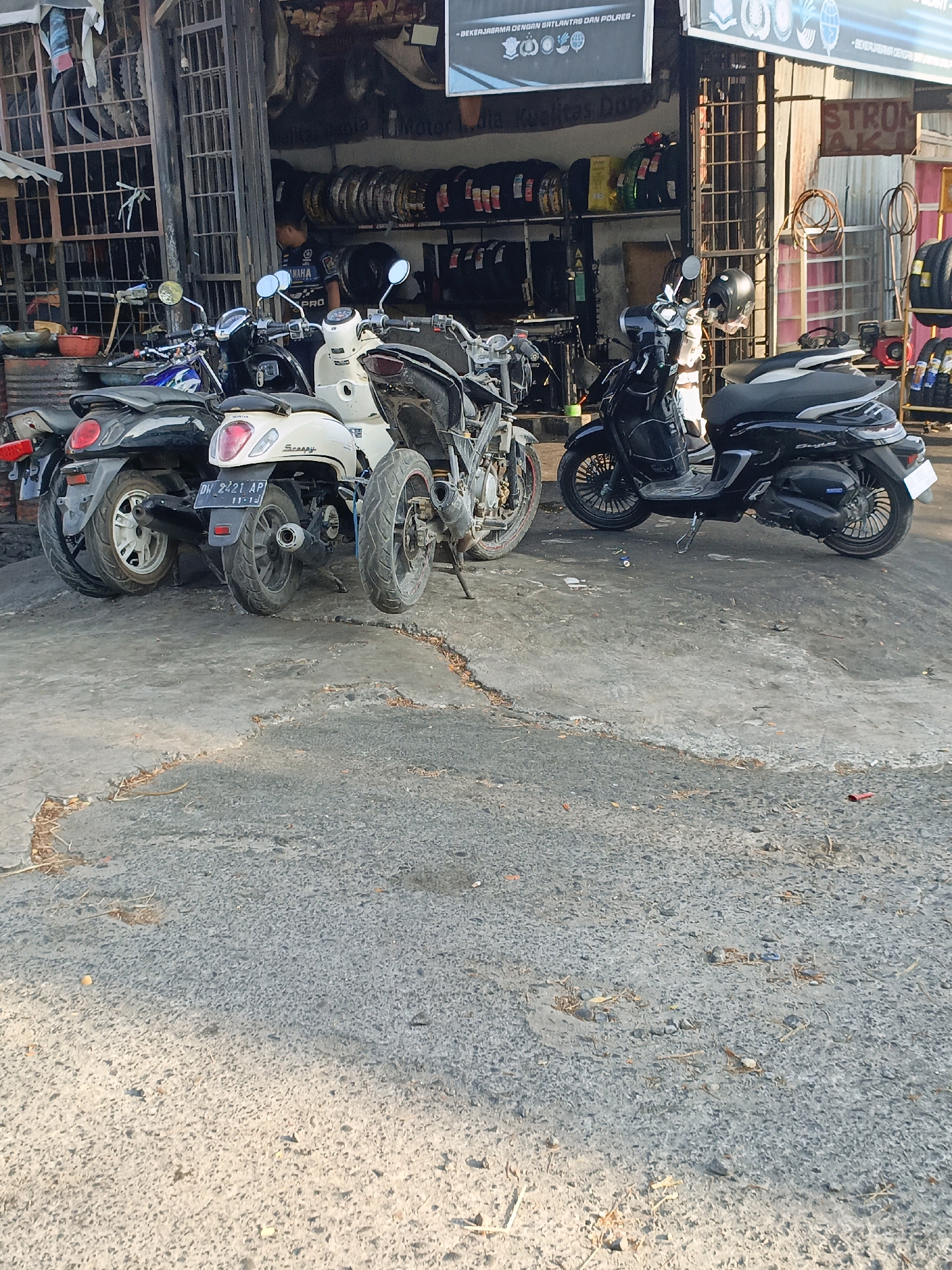 This is a medium shot of a motorcycle repair shop in Bulukumba, Indonesia. Several motorcycles are parked in front of the shop, which has a corrugated metal facade. The motorcycles are of various models and colors, including a white scooter with a black seat and a black scooter with a black seat. A man wearing a blue shirt is standing inside the shop, possibly working. The shop appears to be stocked with tires and other automotive parts. The ground in front of the shop is paved with asphalt and shows signs of wear and tear. The overall impression is that of a busy, functional repair shop in a typical Indonesian city.