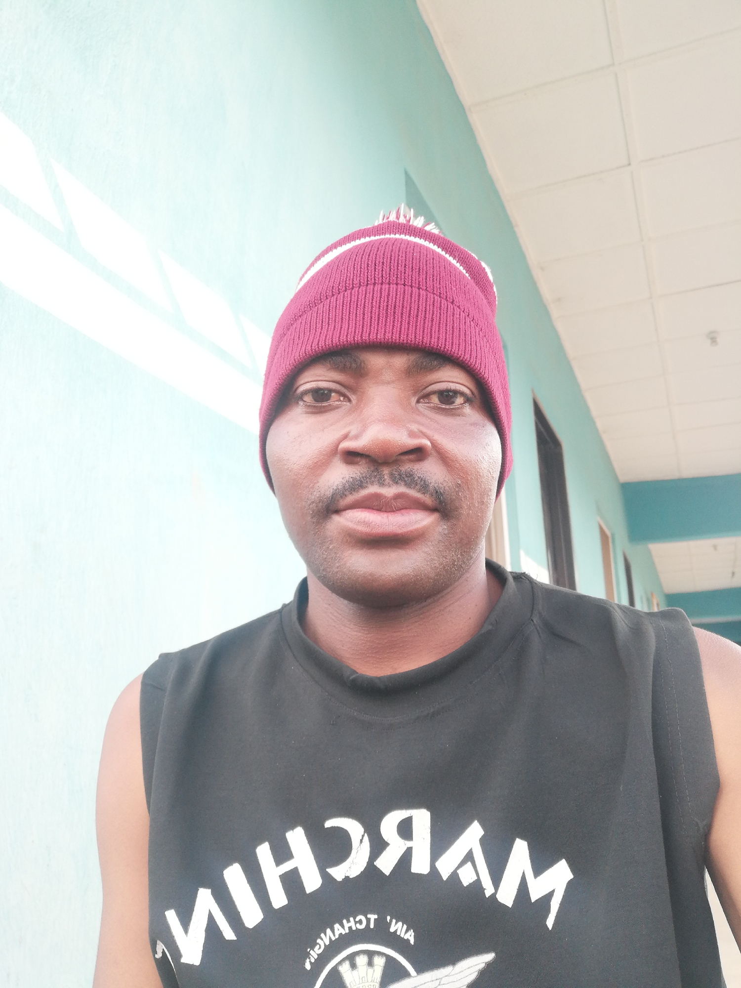 A medium shot captures a man wearing a maroon knitted beanie with a white stripe and a black sleeveless t-shirt. He has a mustache and is looking directly at the camera. The background is a light blue wall with white geometric patterns and a ceiling with white tiles. The scene appears to be an outdoor walkway or corridor, potentially part of a building in Ajasse Ipo, Nigeria.