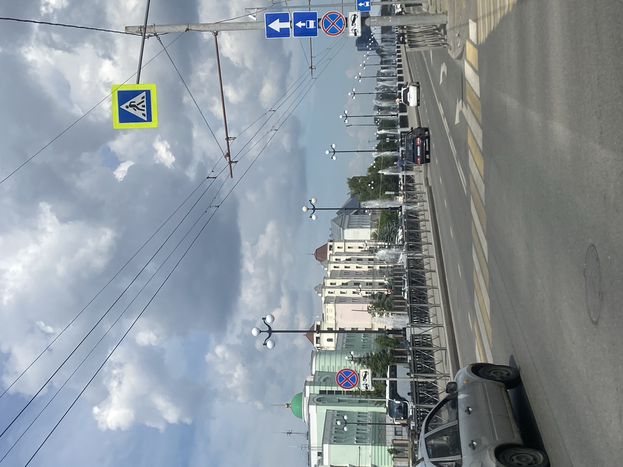 The image captures a street scene in Kazan, Russia, likely during daytime with cloudy skies. In the foreground, a silver sedan is parked on the right side of a pedestrian crossing marked by yellow and white stripes. The road has multiple lanes and appears to be a busy thoroughfare. On the right side of the road, a utility pole displays several traffic signs: a blue sign with a white arrow pointing up, a similar sign with a bus symbol and an upward arrow, a circular red and blue sign indicating no stopping or parking, and a sign depicting a tow truck. Further down the street, several cars are visible, including a dark sedan and a white car.
On the left side of the road, a black metal railing separates the street from a sidewalk or promenade. Beyond the railing, a row of lampposts with multiple globes illuminates the area. In the background, a line of buildings with distinctive architecture, including a building with a green dome and pale green facades, are visible. Fountains are operating, with water jets creating a misty effect in front of the buildings. The sky is filled with dramatic, dark clouds, suggesting an impending change in weather. Overhead, power lines crisscross the sky. The overall impression is a snapshot of an urban environment with its characteristic infrastructure and elements of city life.